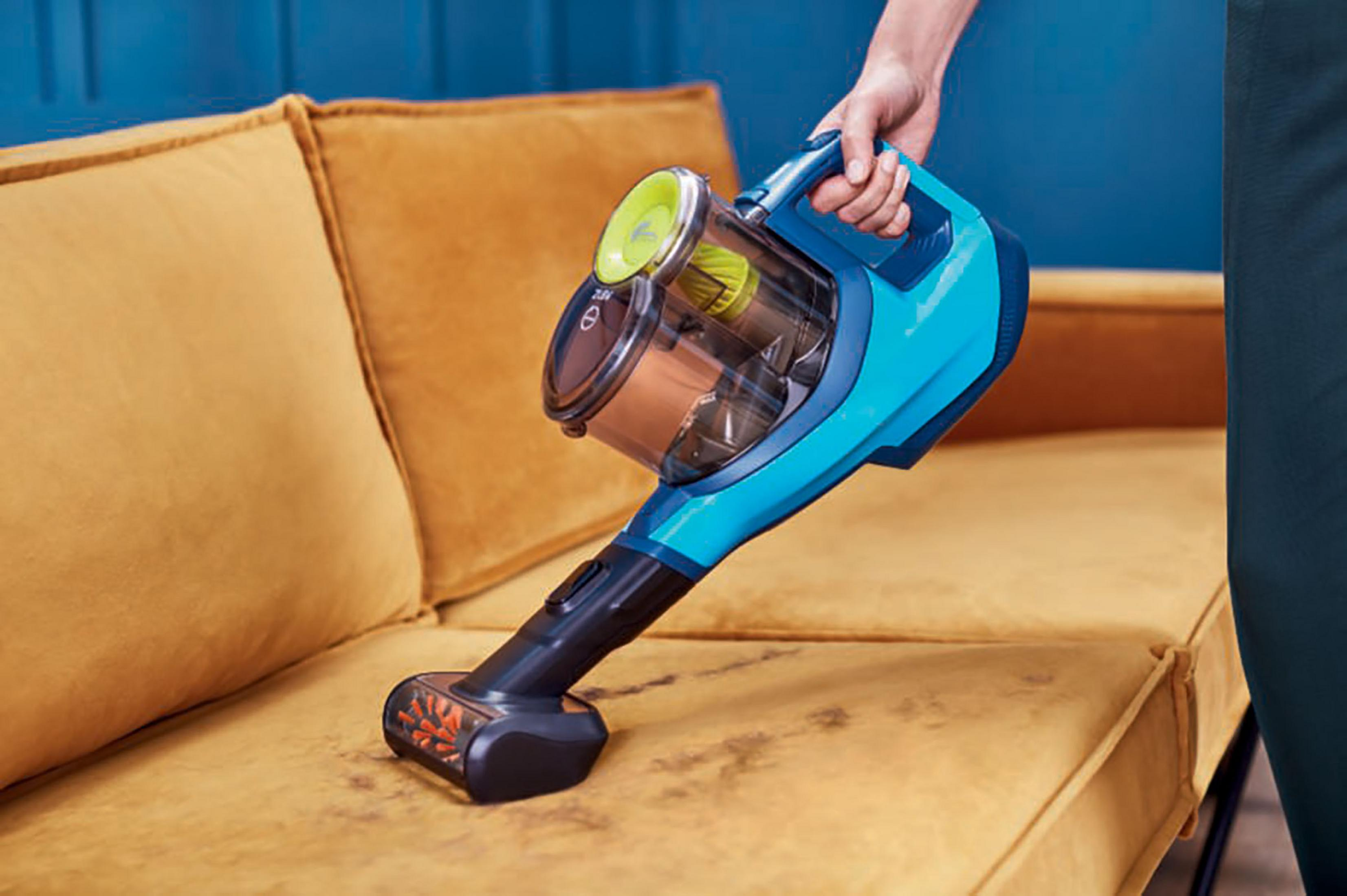 Ręka używająca niebieskiego odkurzacza na żółtej sofie z plamami brudu.