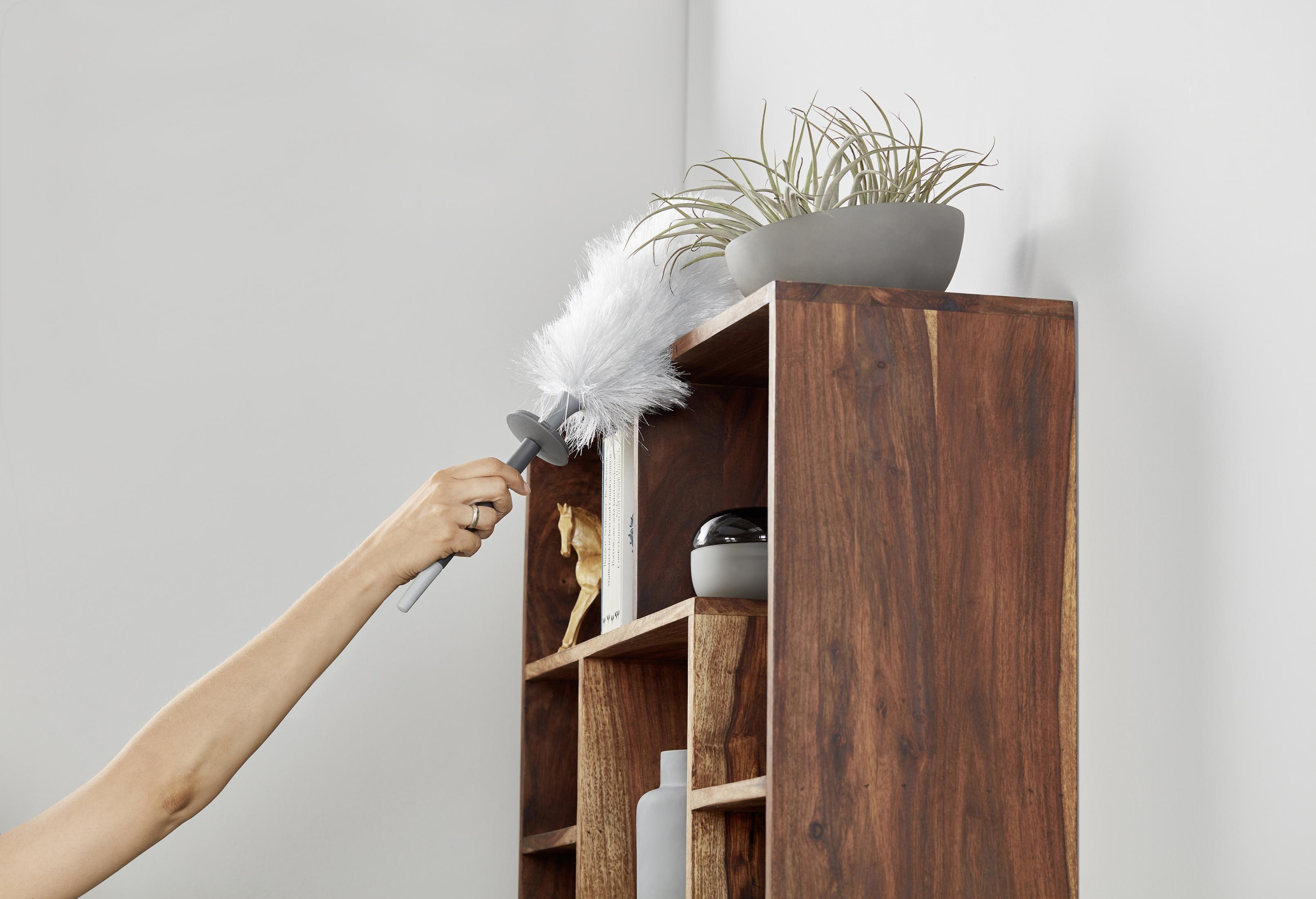 Una persona desempolvando una estantería de madera con un plumero, en un rincón.