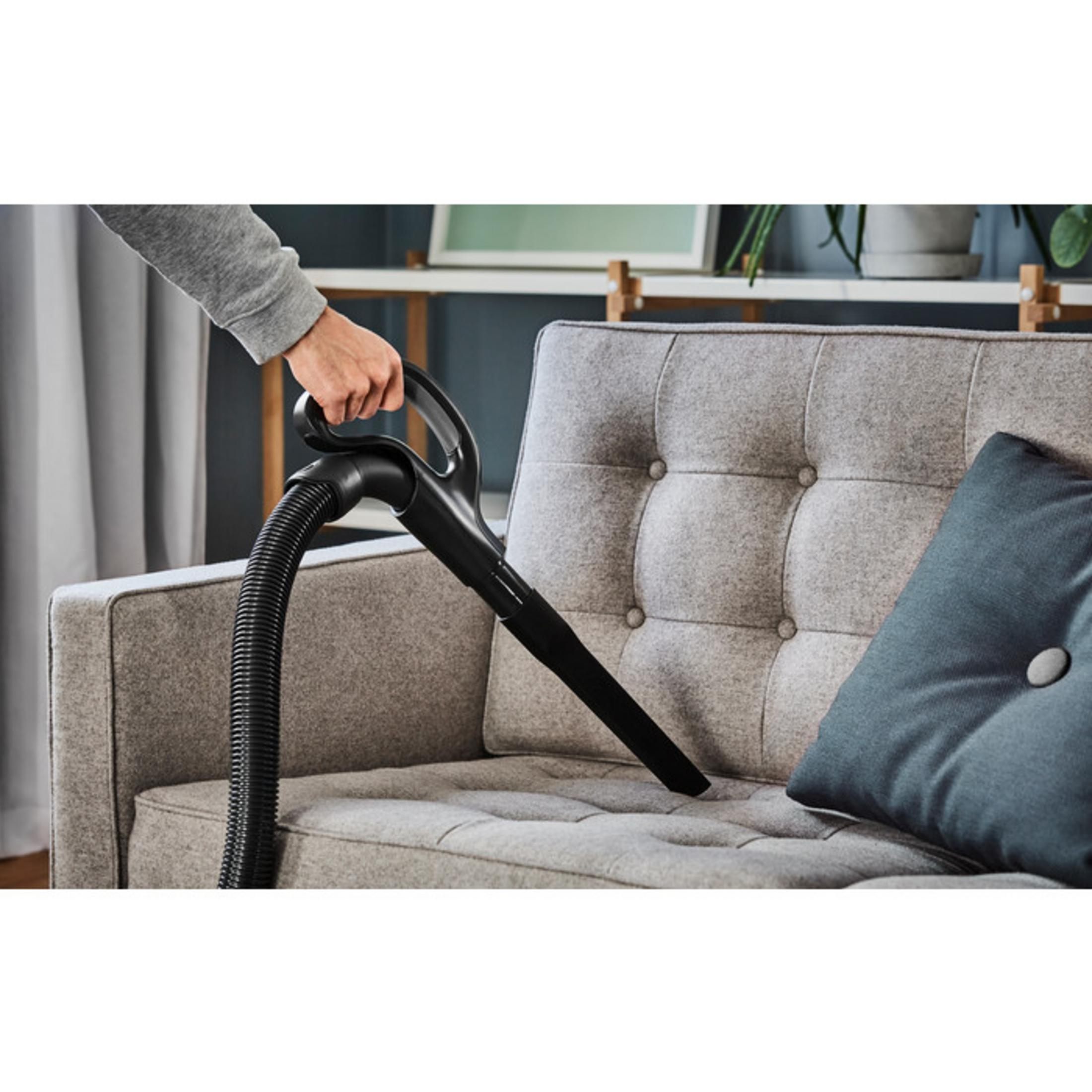 Hand einer Person reinigt mit einem Staubsauger ein graues Sofa. Schwarzer Schlauch und Düse sichtbar.