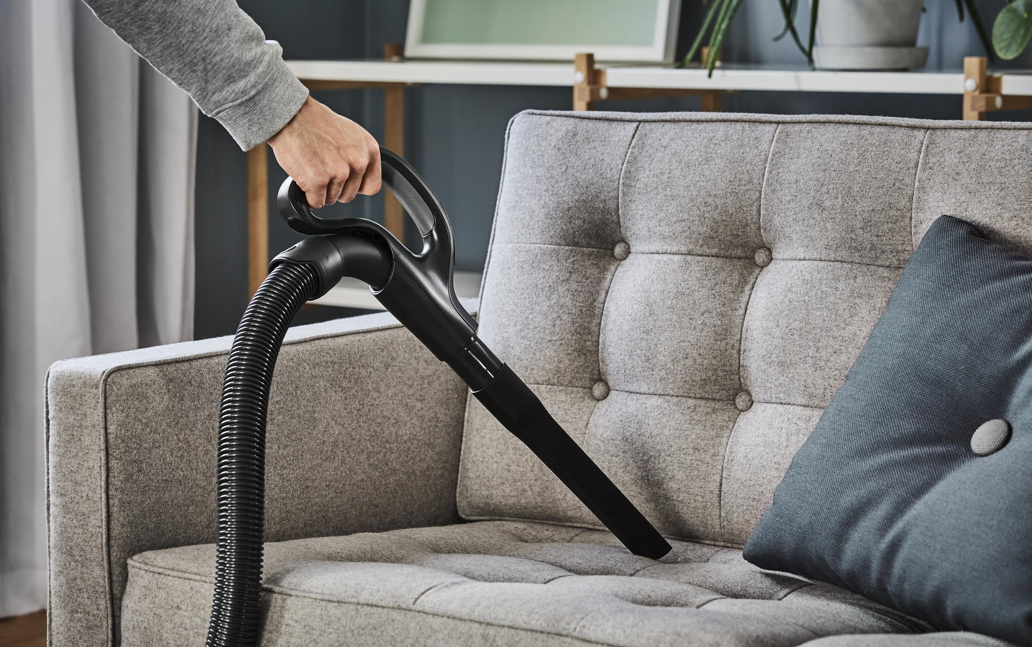 Eine Person saugt ein graues Sofa mit einem schwarzen Staubsauger ab und konzentriert sich auf die Kissen.