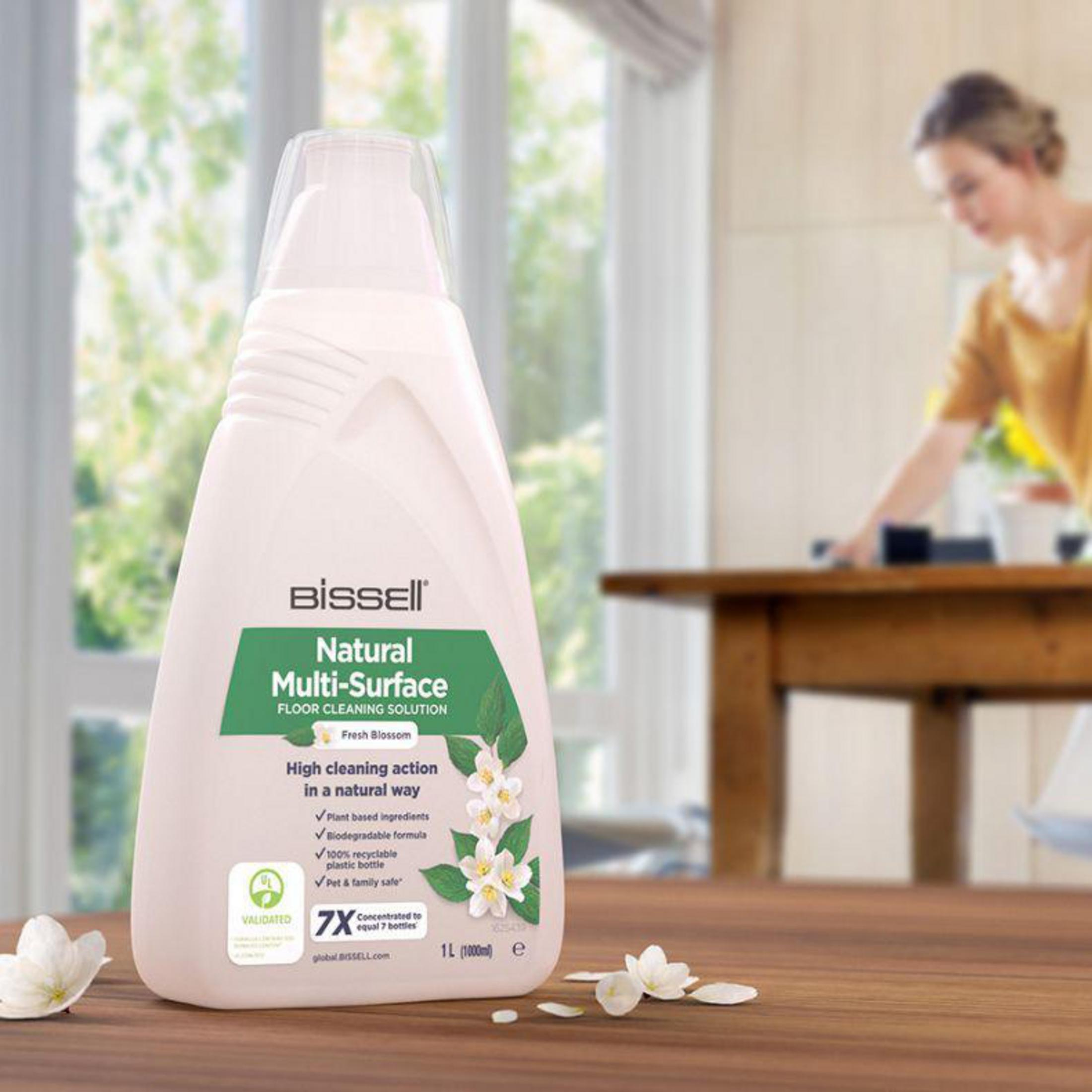 Een vrouw op de achtergrond en een fles Bissell vloerreiniger op een houten tafel.