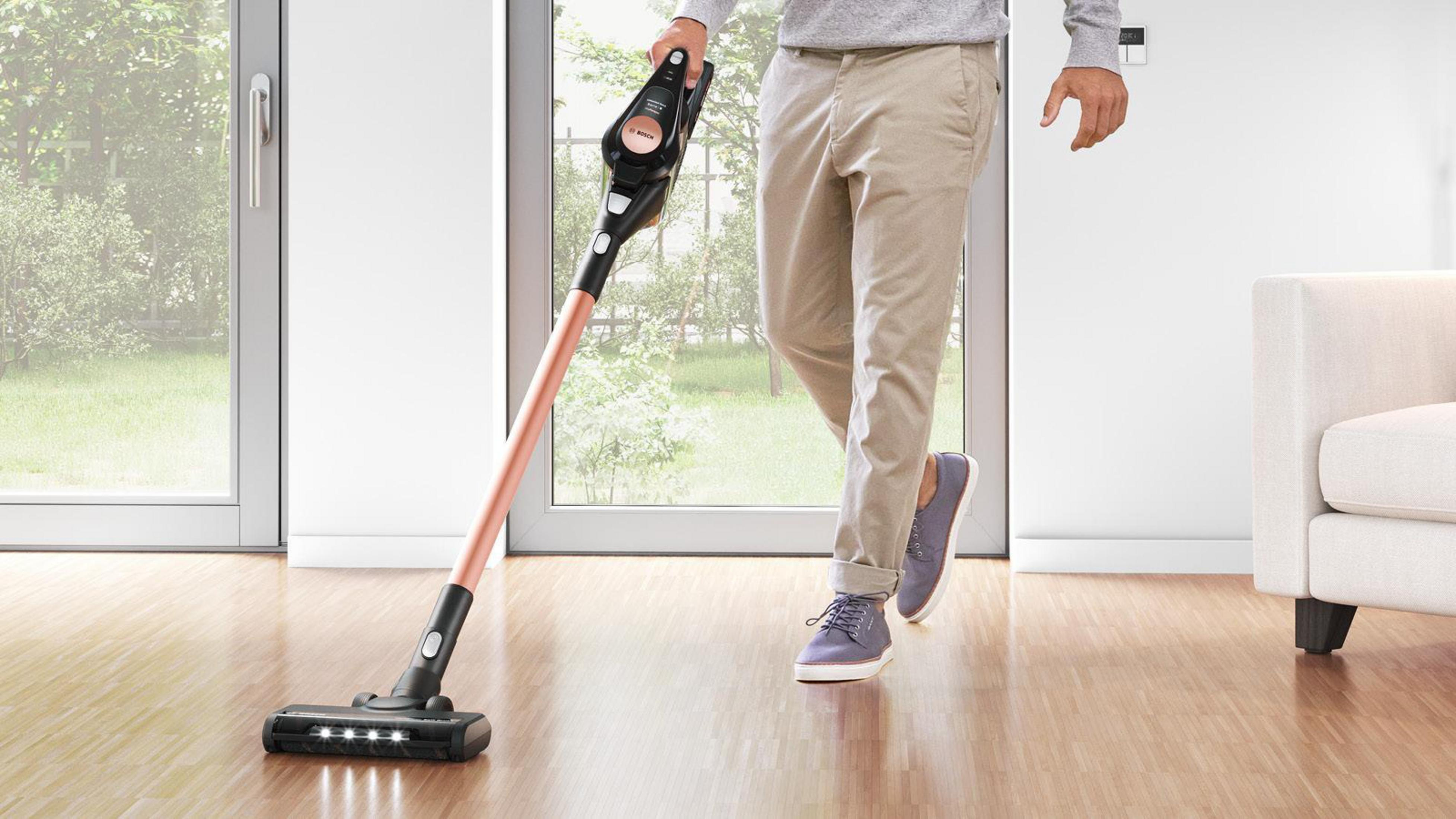 Mężczyzna używa różowego złotego odkurzacza na drewnianej podłodze. W tle okno i sofa.