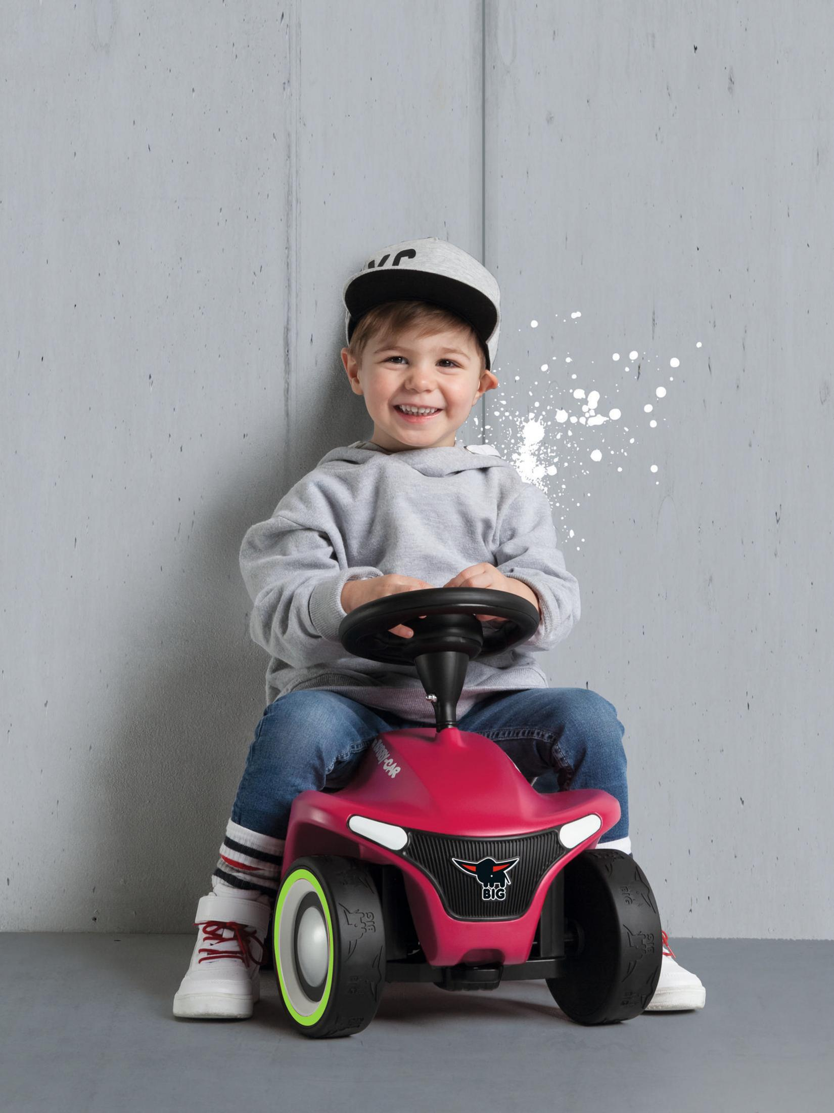 Un niño sentado en un coche de juguete rosa, sonriendo. Fondo de pared gris.
