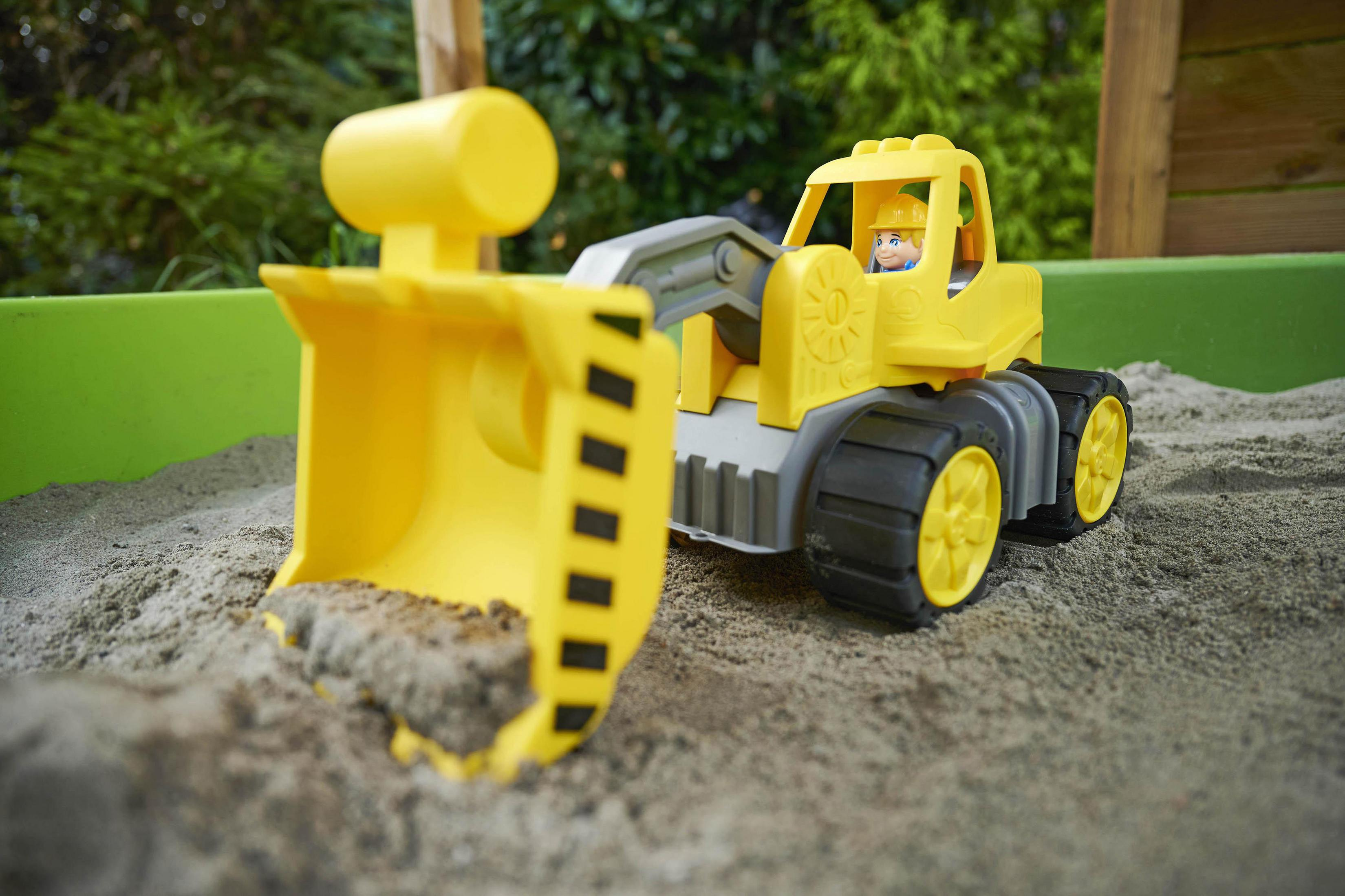 Żółty zabawkowy buldożer z piaskiem w łyżce. Dziecko siedzi na miejscu kierowcy. Zielone tło.