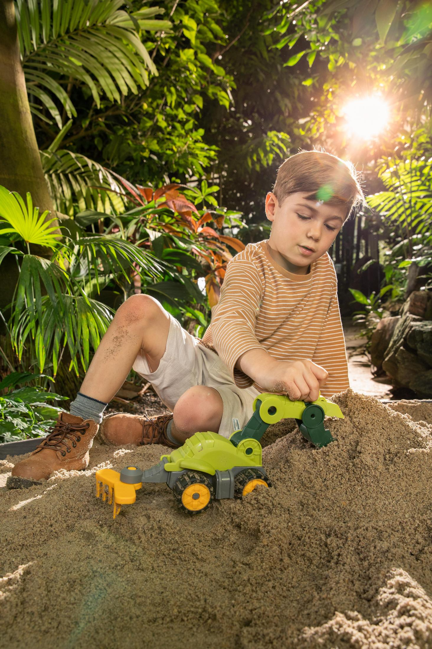 Ein Junge spielt mit einem Spielzeugbagger im Sand. Grünes und gelbes Spielzeug. Grünes Gemüse dahinter.