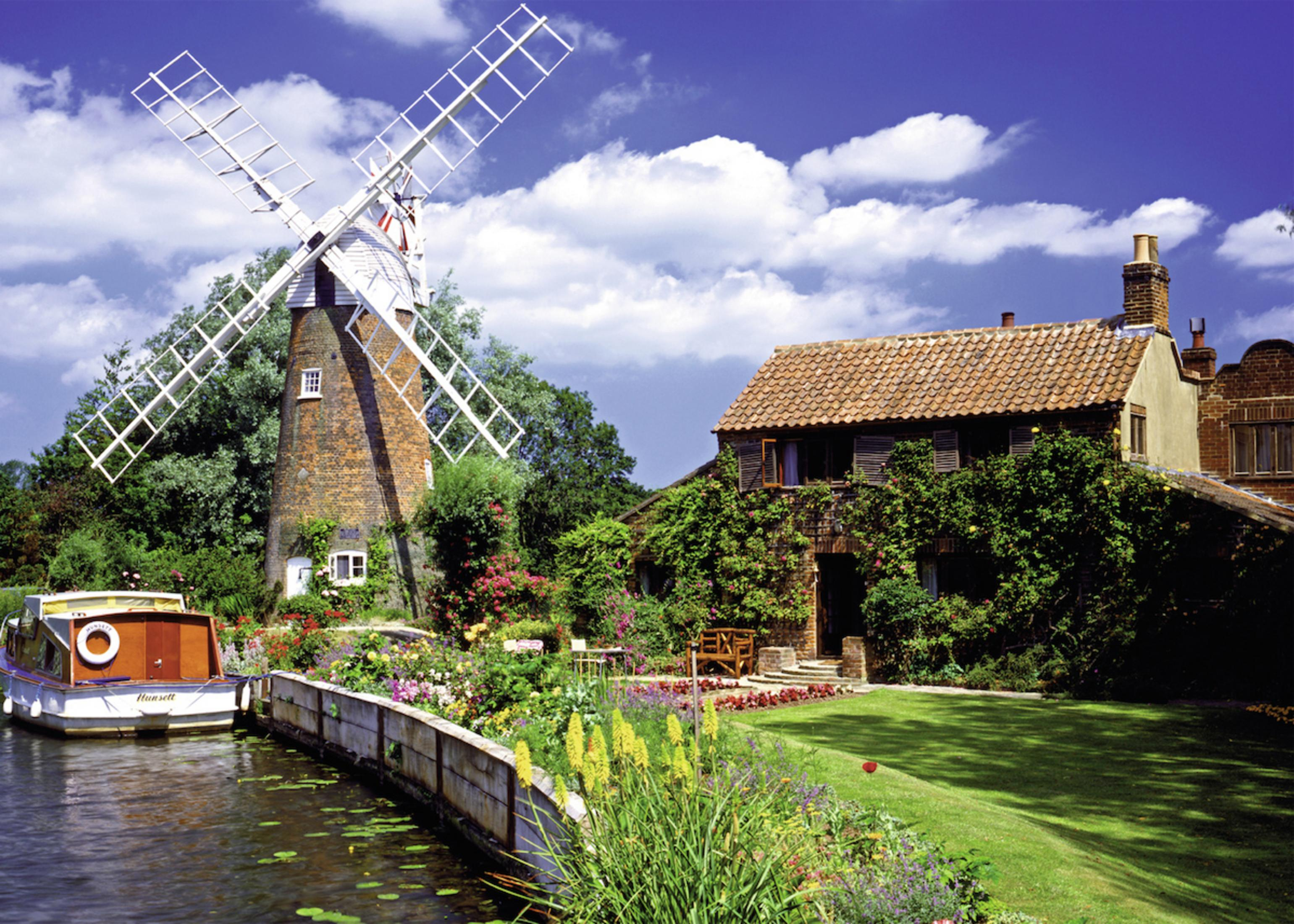 Eine Windmühle, ein Haus und ein Boot auf einem Fluss. Blumen und Blätter umgeben die Gebäude. Blauer Himmel mit Wolken.