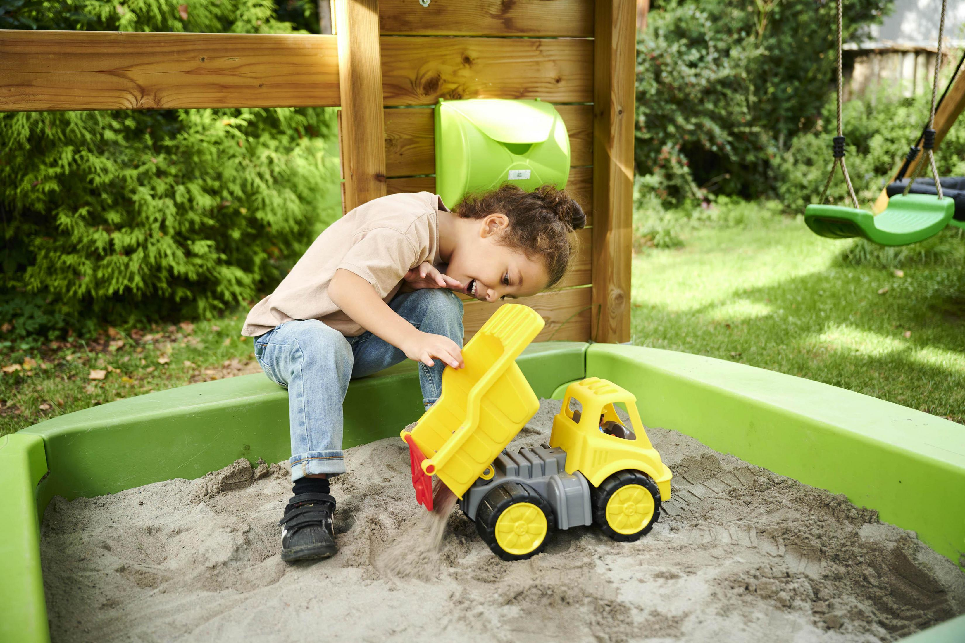 Ein Kind spielt in einem Sandkasten mit einem gelben Spielzeug-Lastwagen. Grüner Sandkasten umgibt.