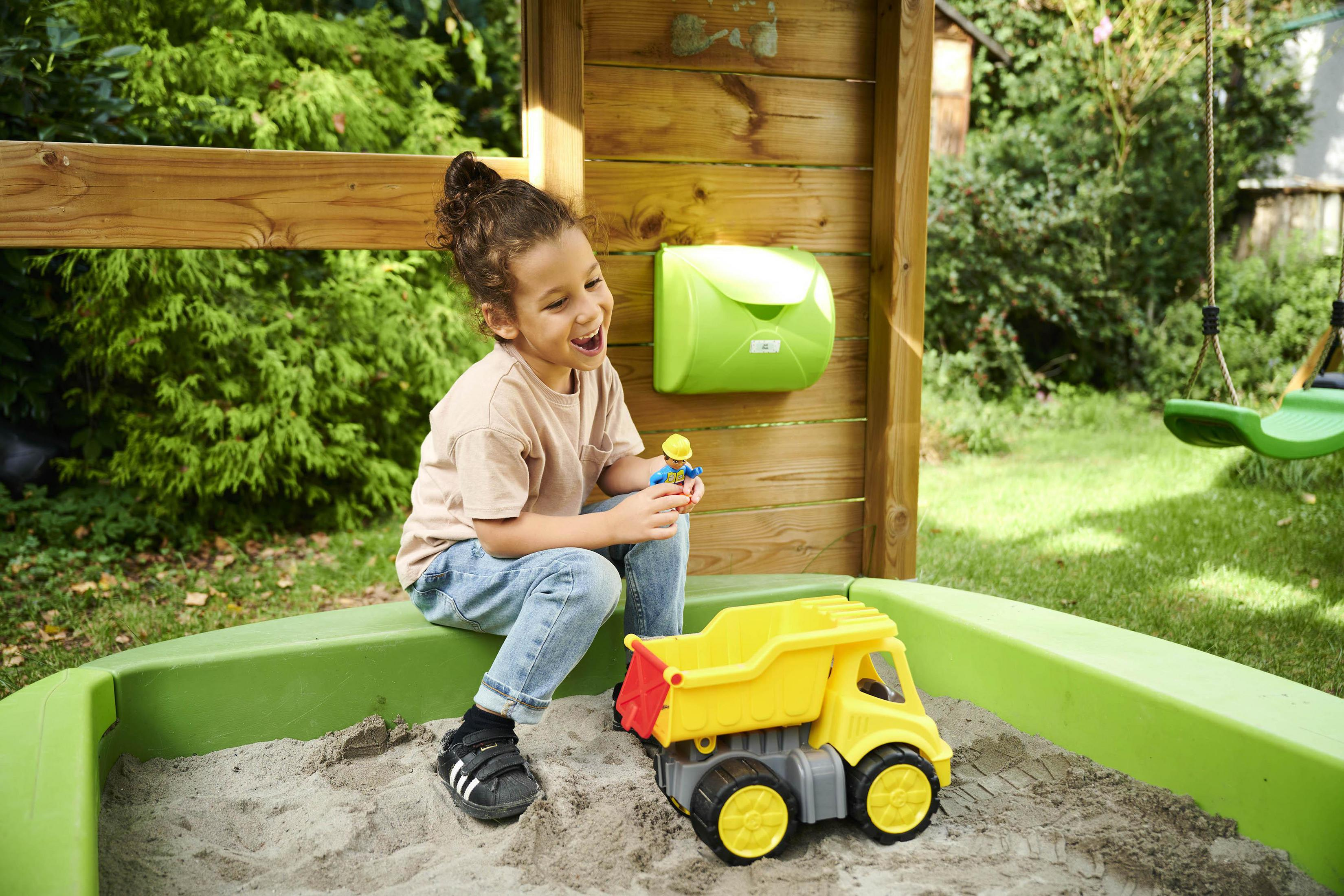 Ein junges Kind lacht in einem Sandkasten und spielt mit einem gelben Spielzeug-LKW und einer Figur.