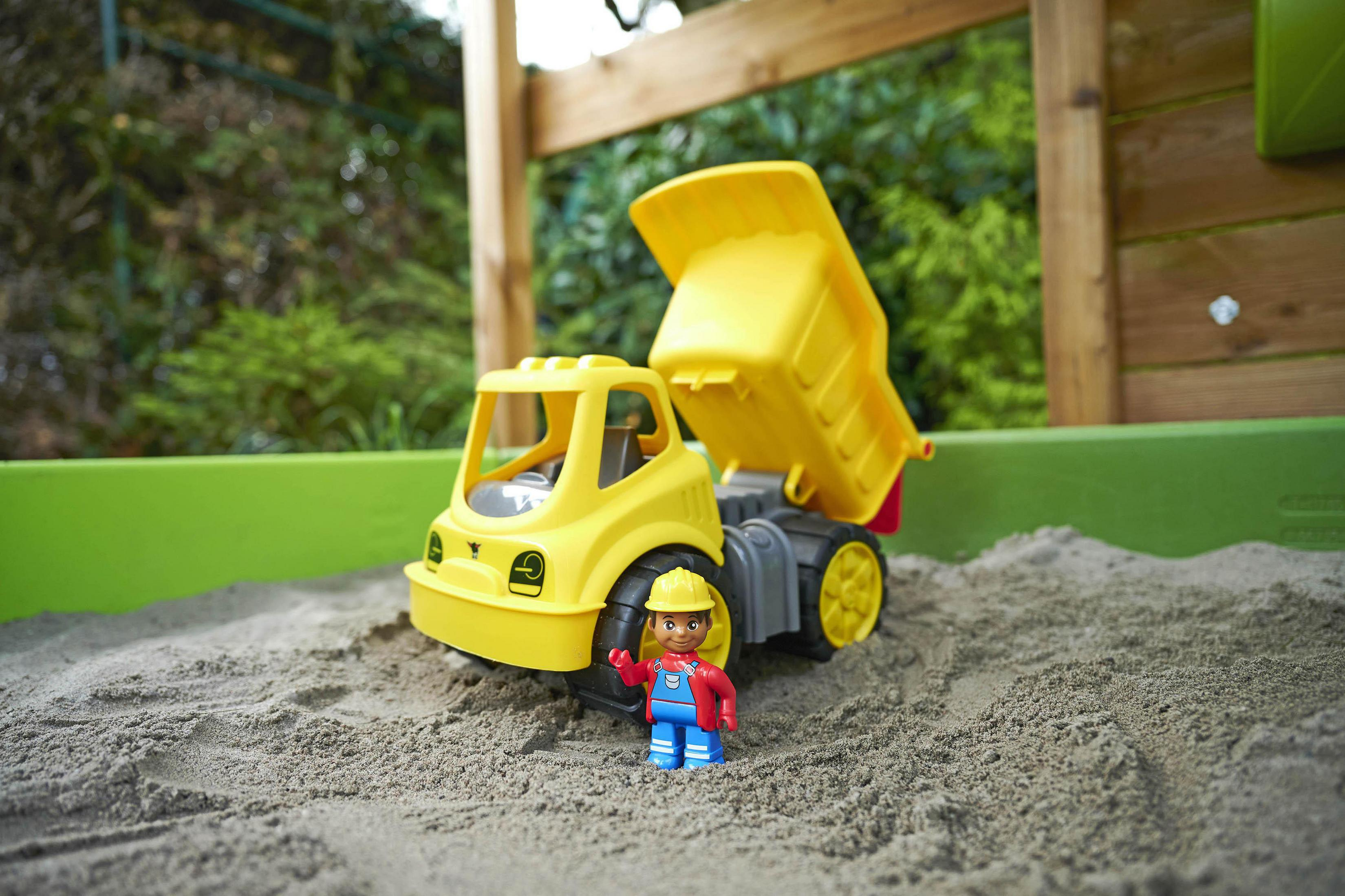Ein gelber Spielzeug-Muldenkipper ist mit einer Lego-Figur eines Bauarbeiters im Sandkasten.