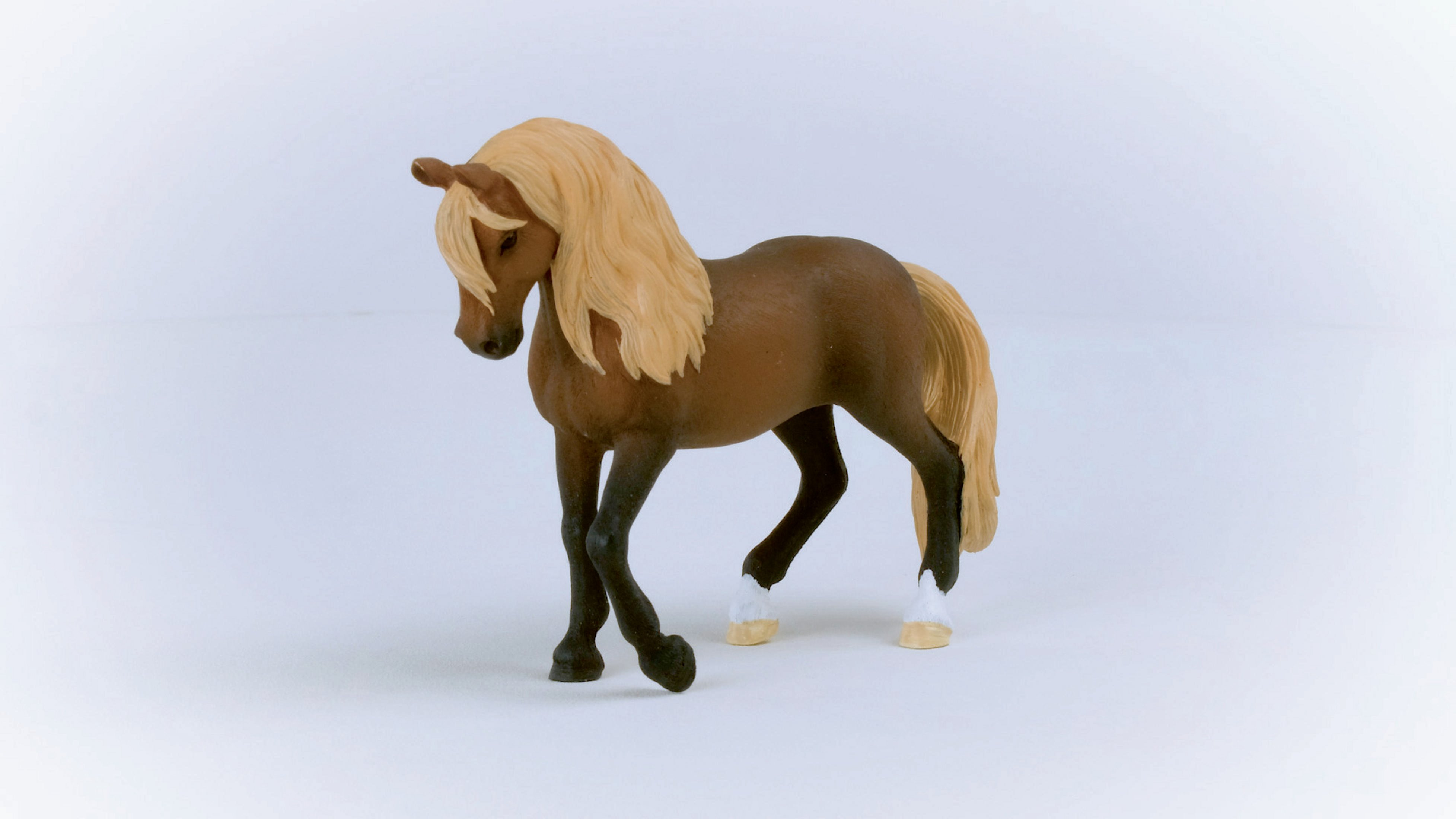 Brązowa zabawka konia z blond grzywą, stojąca na białej powierzchni. Tło jest rozmyte.