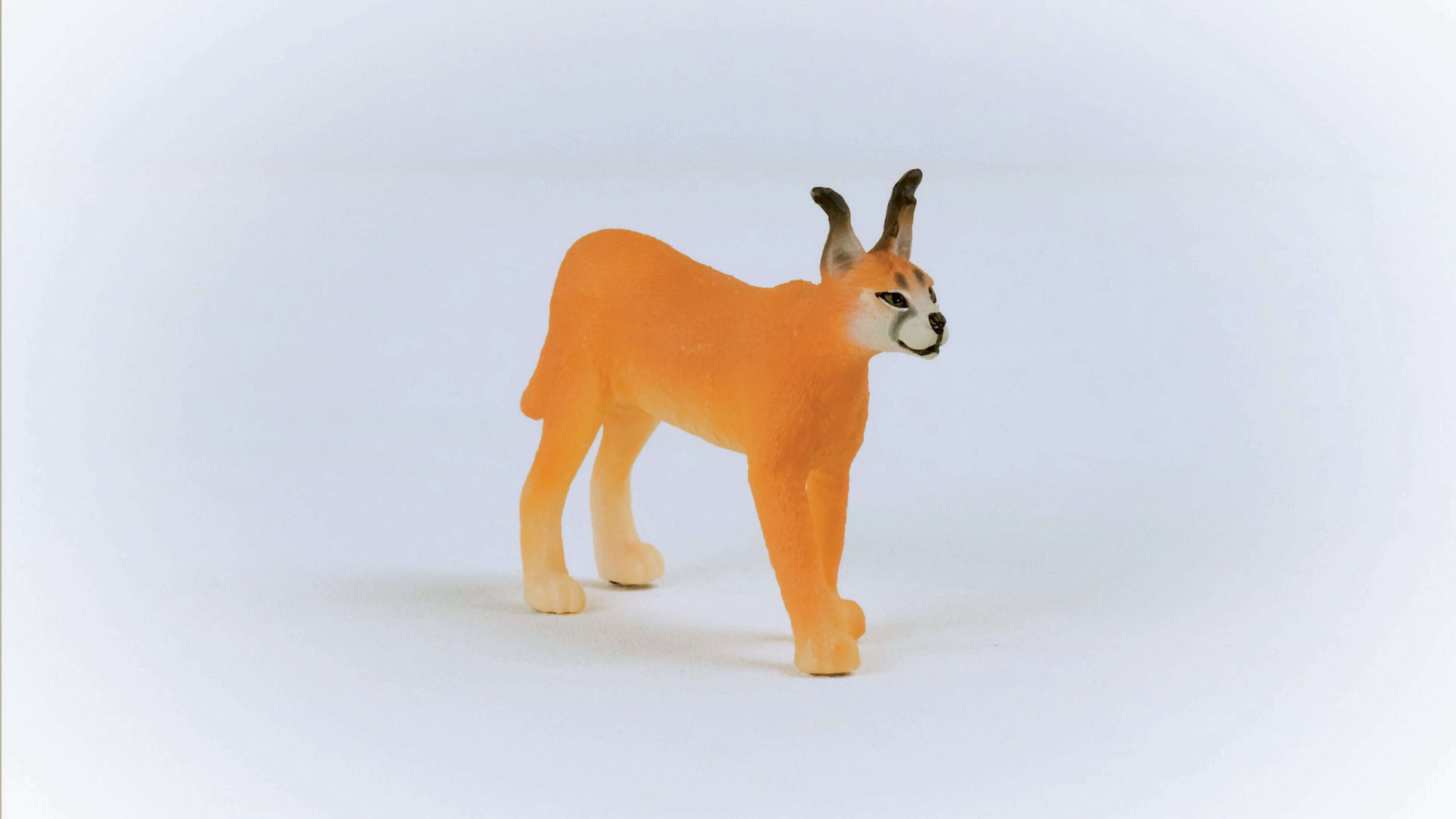 Pomarańczowa figurka karakala stoi na białej powierzchni. Karakal ma uszy.