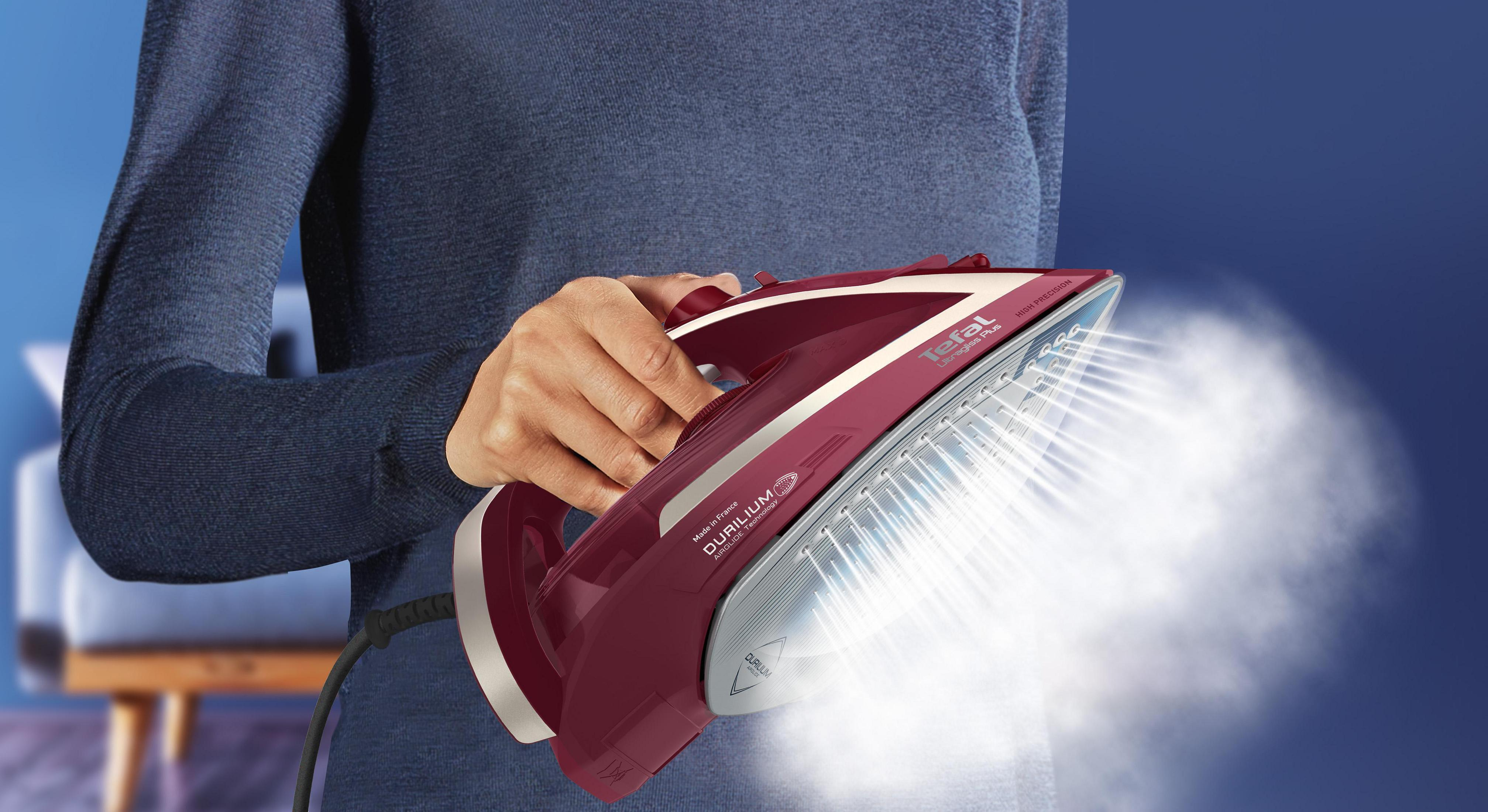 Osoba prasująca żelazkiem parowym w kolorze bordowym, widoczna para, niebieskie tło i krzesło.