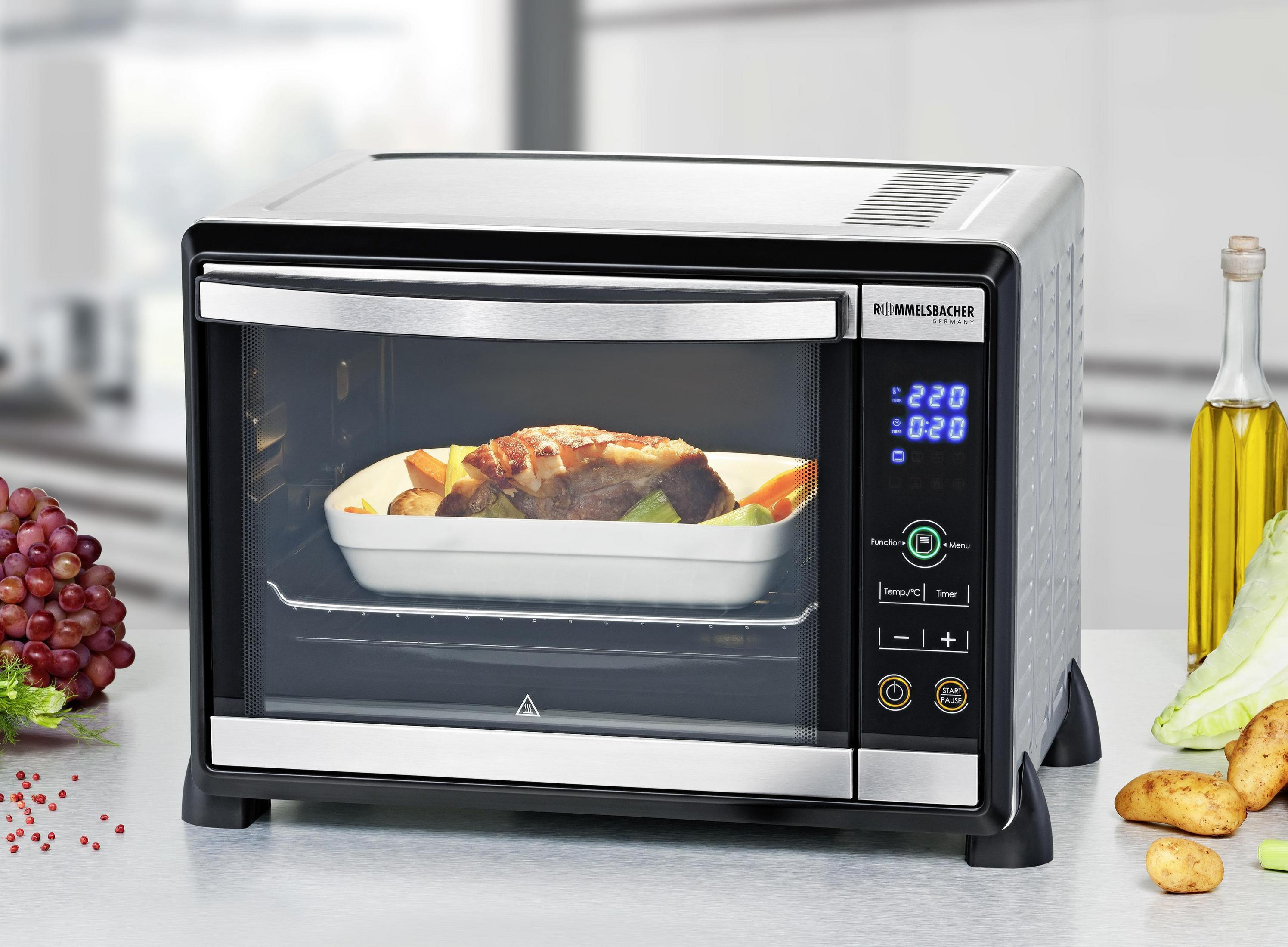 Czarny i srebrny piekarnik z jedzeniem w środku, warzywami i winogronami w tle.