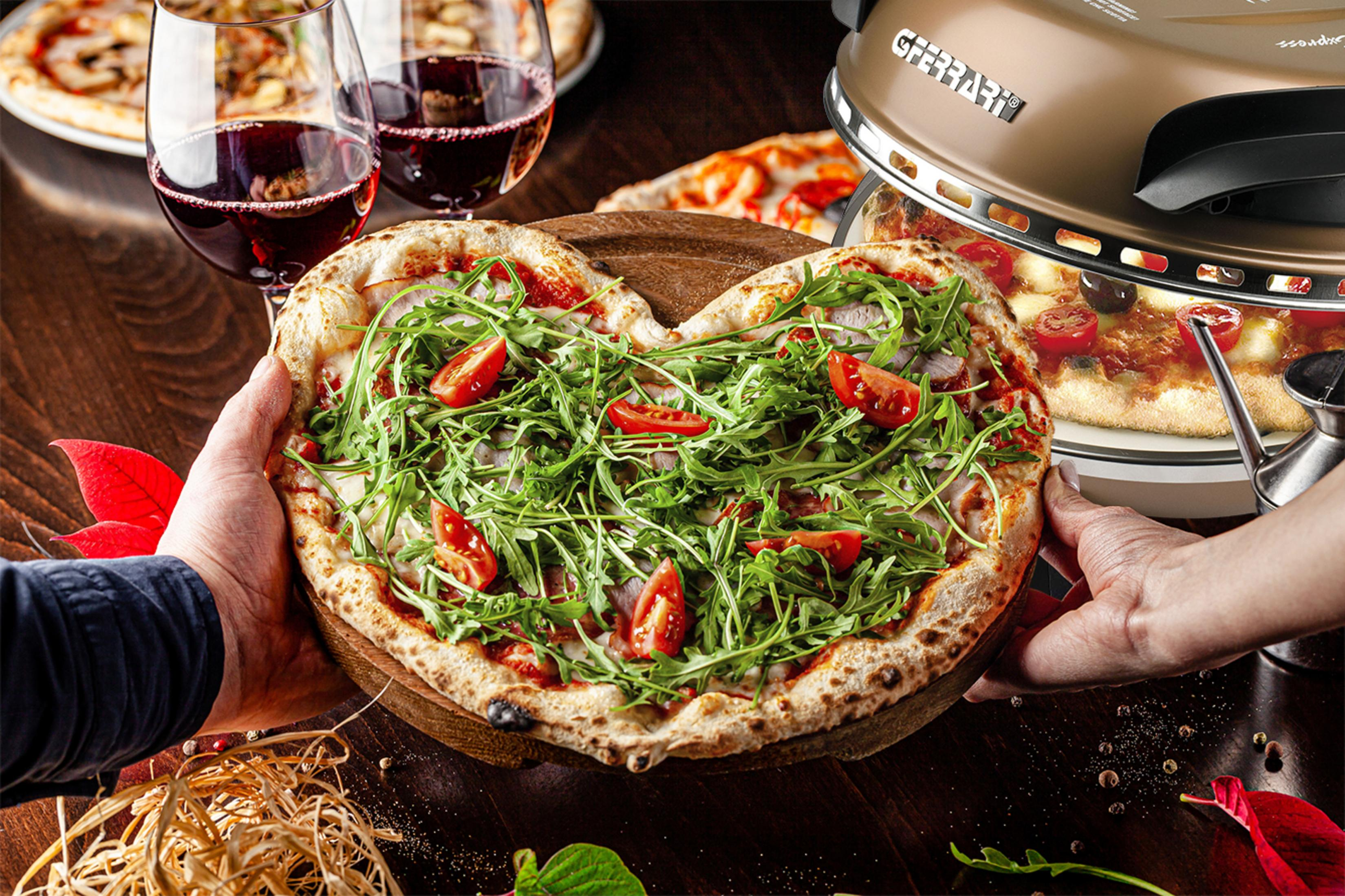 Eine herzförmige Pizza wird gehalten. Weingläser und ein Ofen sind im Hintergrund.