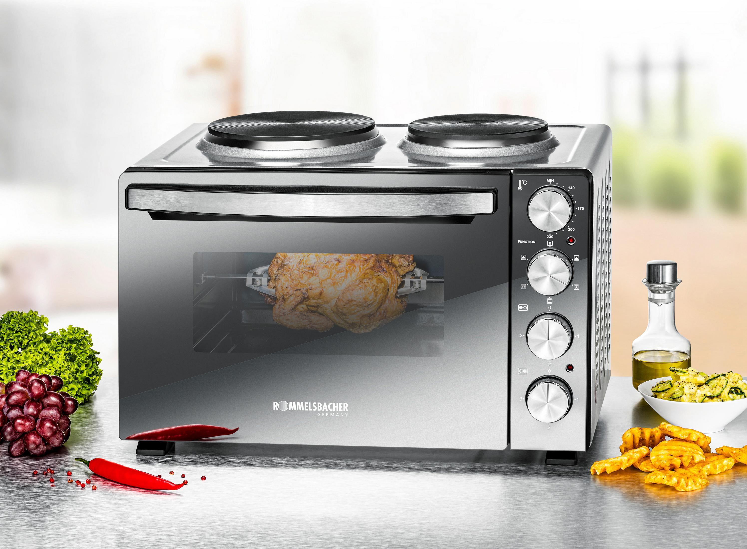 Un forno con due piastre. Un pollo arrostisce all'interno. Uva, insalata e patatine fritte nelle vicinanze.