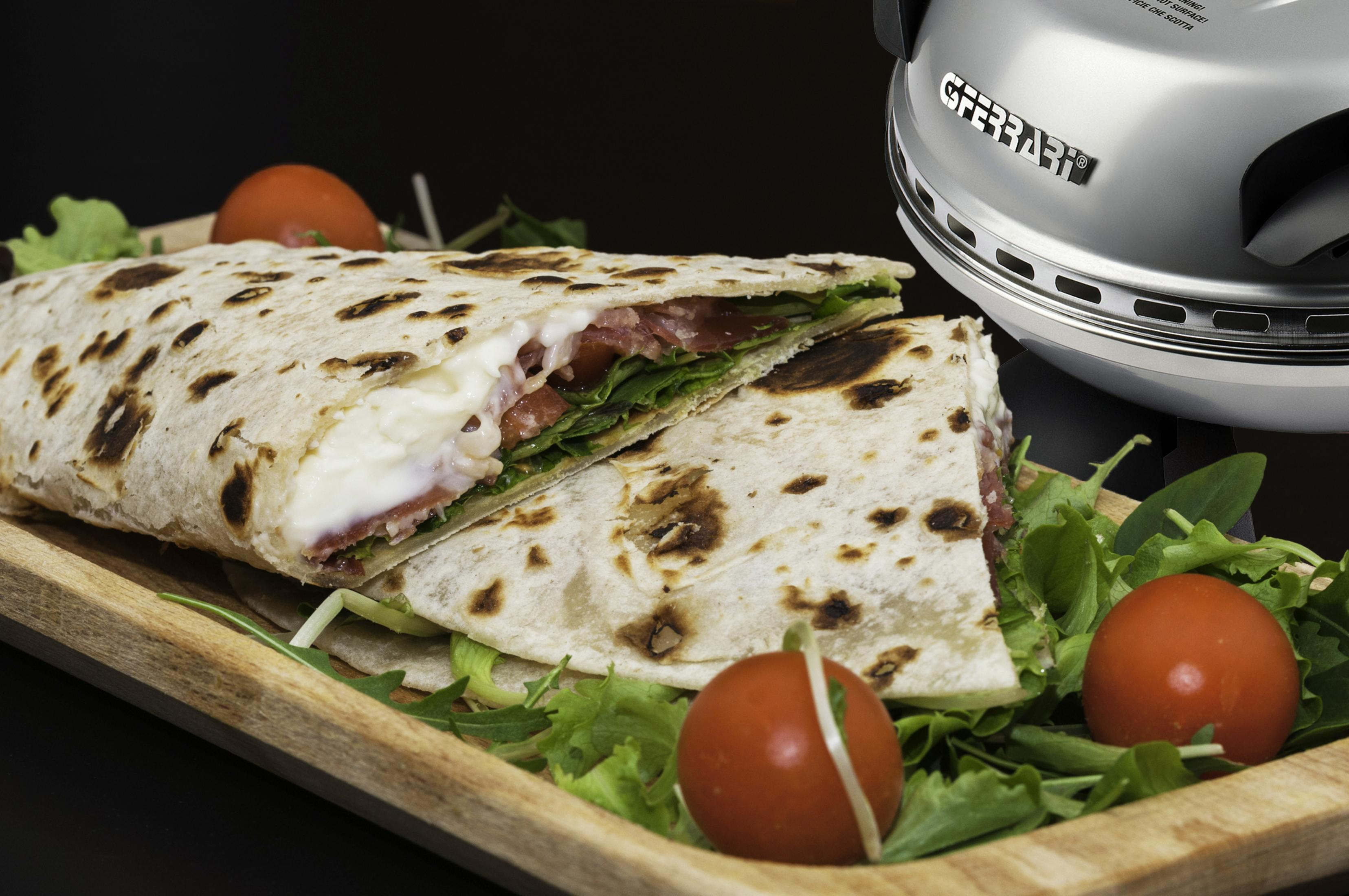 Ein Fladenbrot-Sandwich mit Mozzarella und Tomaten auf einem Holztablett. Ein Pizzaofen ist im Hintergrund.