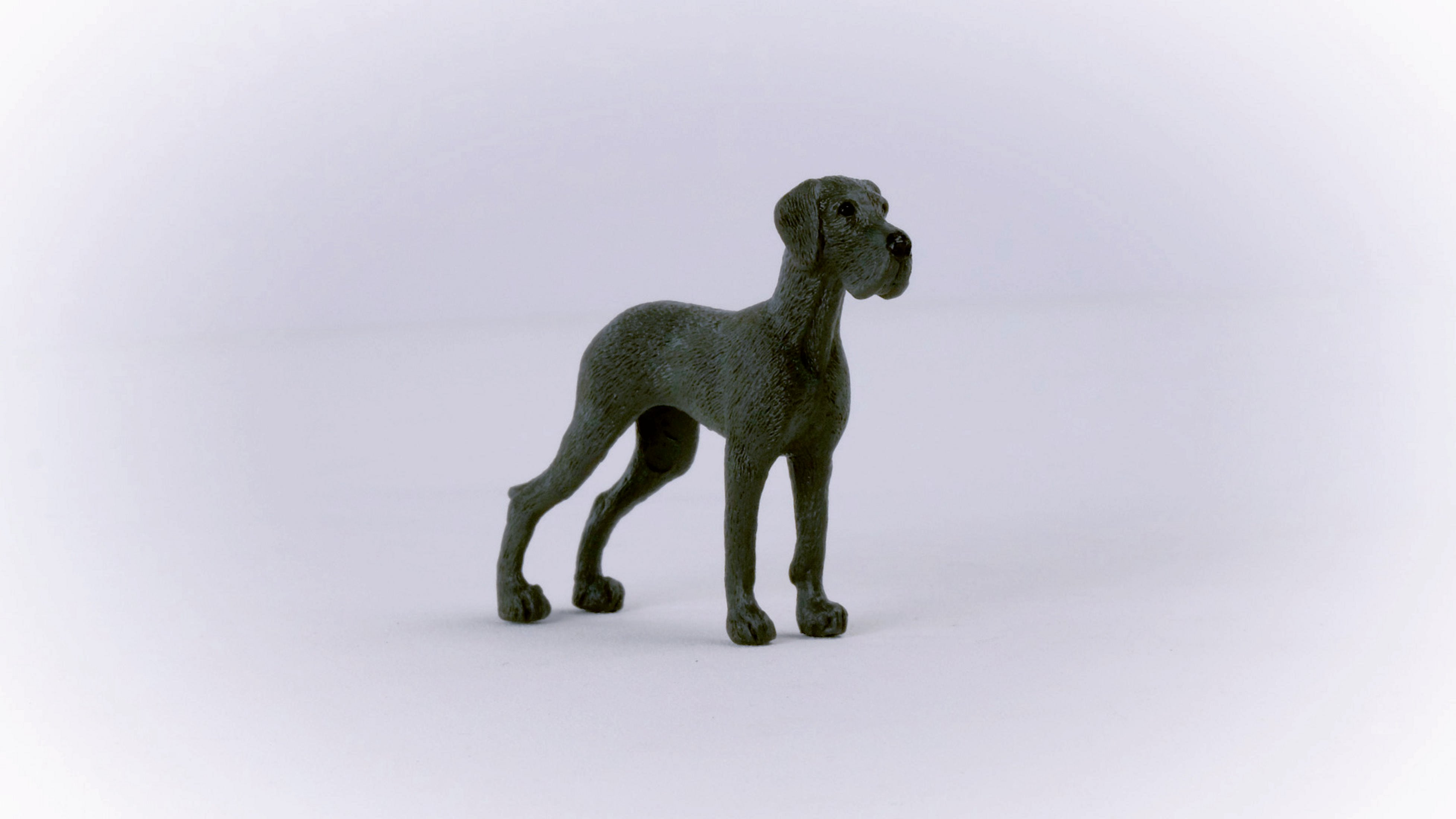 Szara statua doga niemieckiego, stojąca na białej powierzchni. Pies ma smukłą budowę i patrzy w prawo.