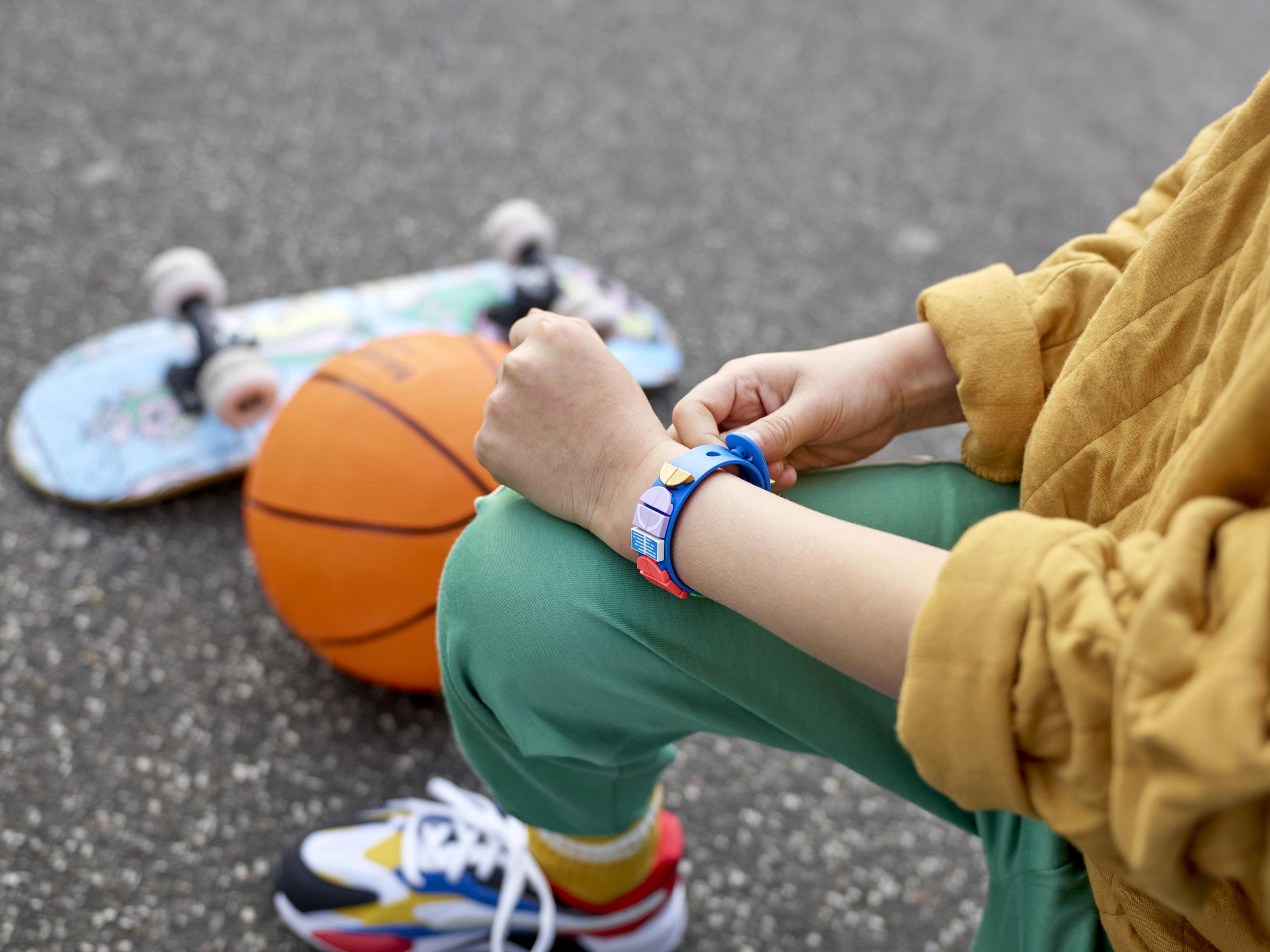 Mavi bir saat takan bir çocuk basketbol, kaykay ve bir ayakkabının yanında oturuyor.
