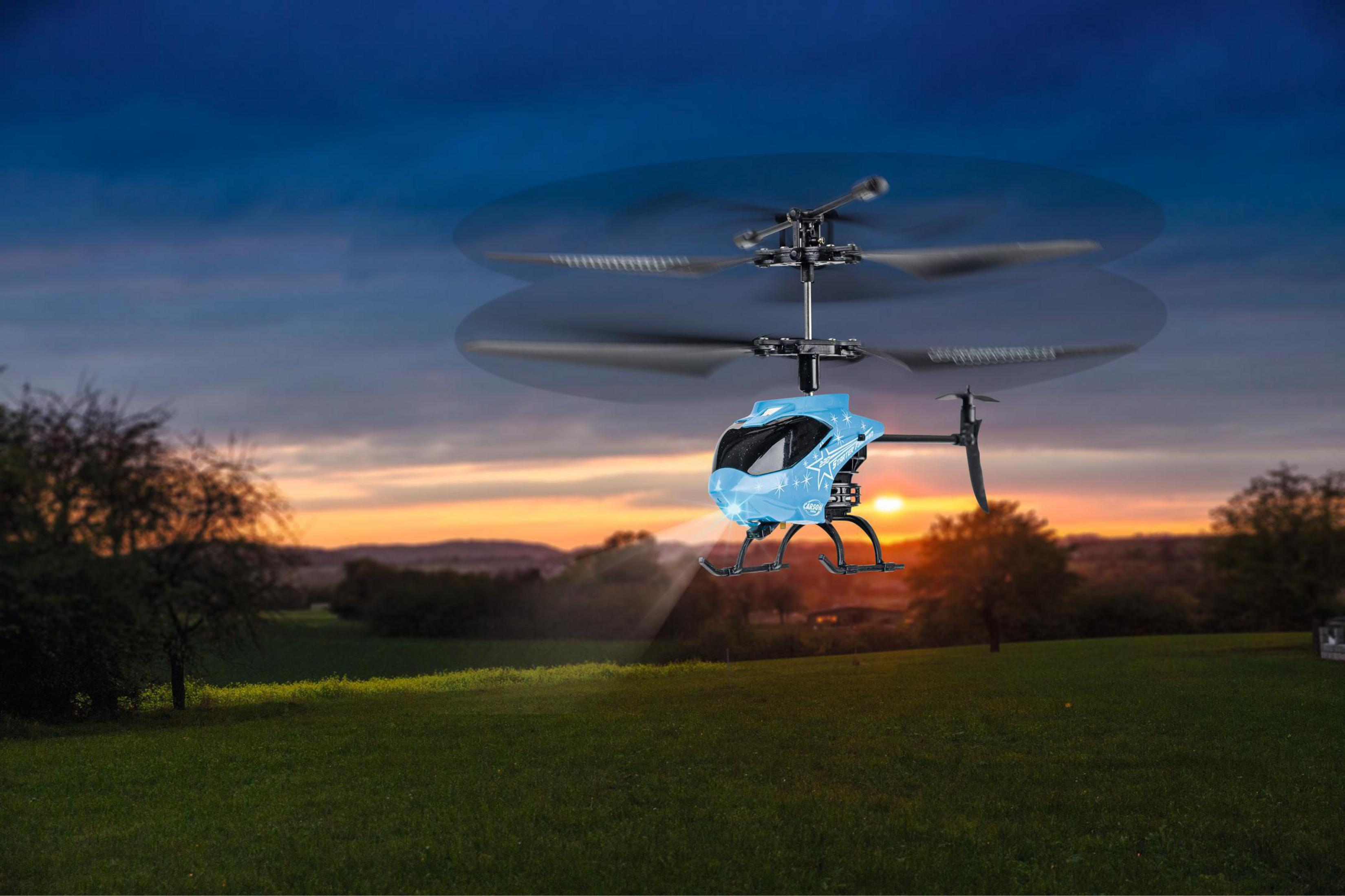 Ein blau-schwarzer Spielzeug-Helikopter, der bei Sonnenuntergang über ein grünes Feld fliegt. Die Rotorblätter sind verschwommen.