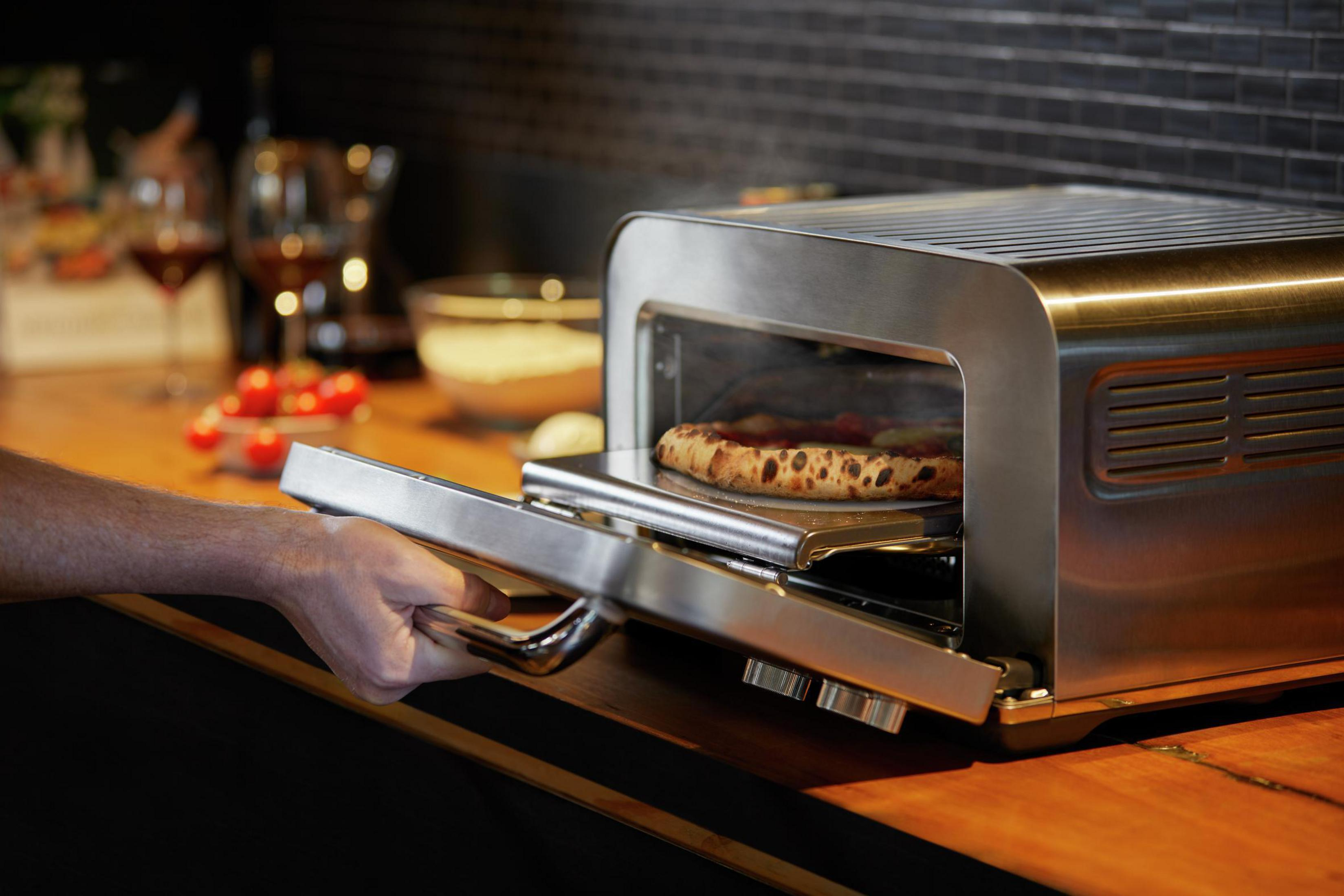 Forno per pizza con una pizza all'interno e una mano che apre la porta, posto su un piano della cucina.