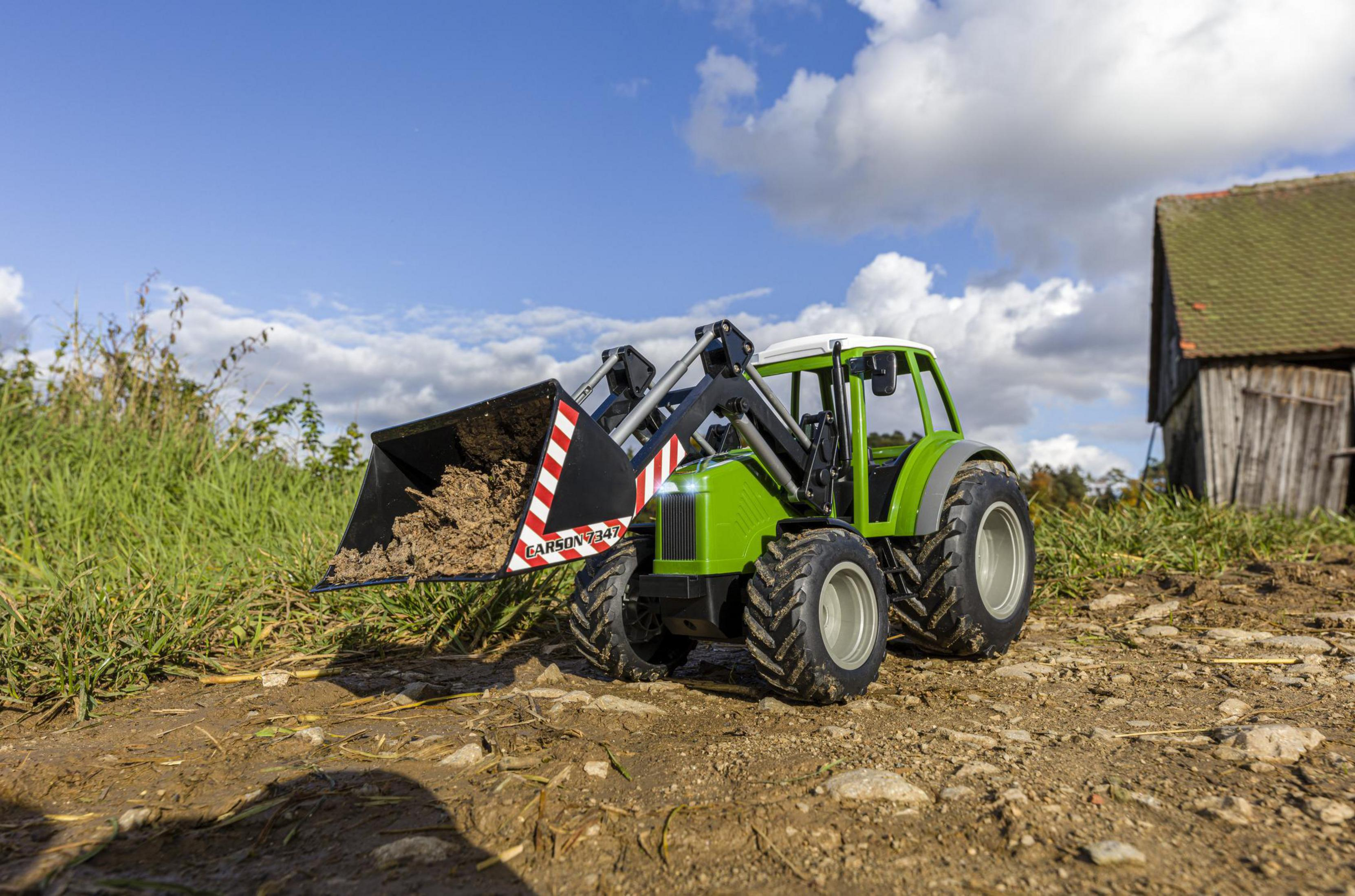 Grüner Spielzeugtraktor mit einem Eimer Erde auf einem Feldweg, mit Feldern und einer Scheune im Hintergrund.