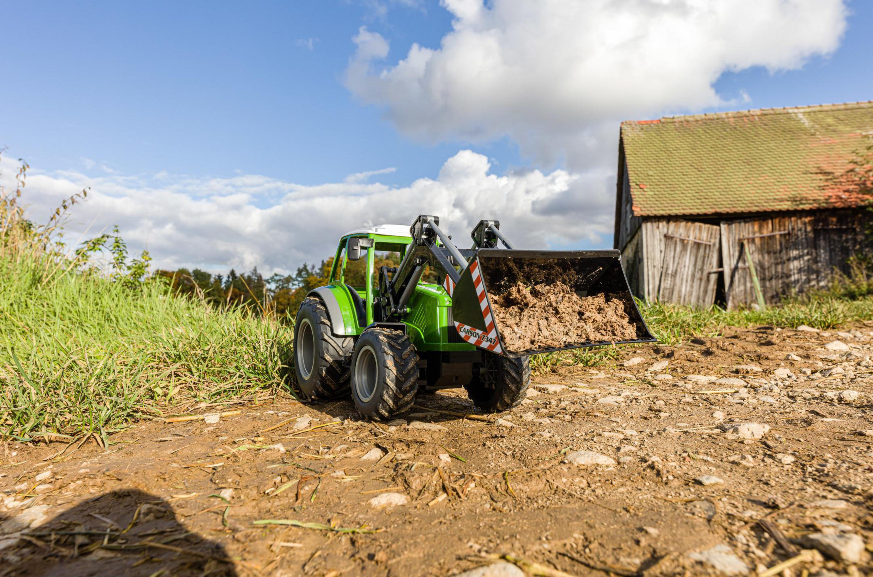 Grüner Traktor lädt Erde, mit Scheune und bewölktem Himmel im Hintergrund.