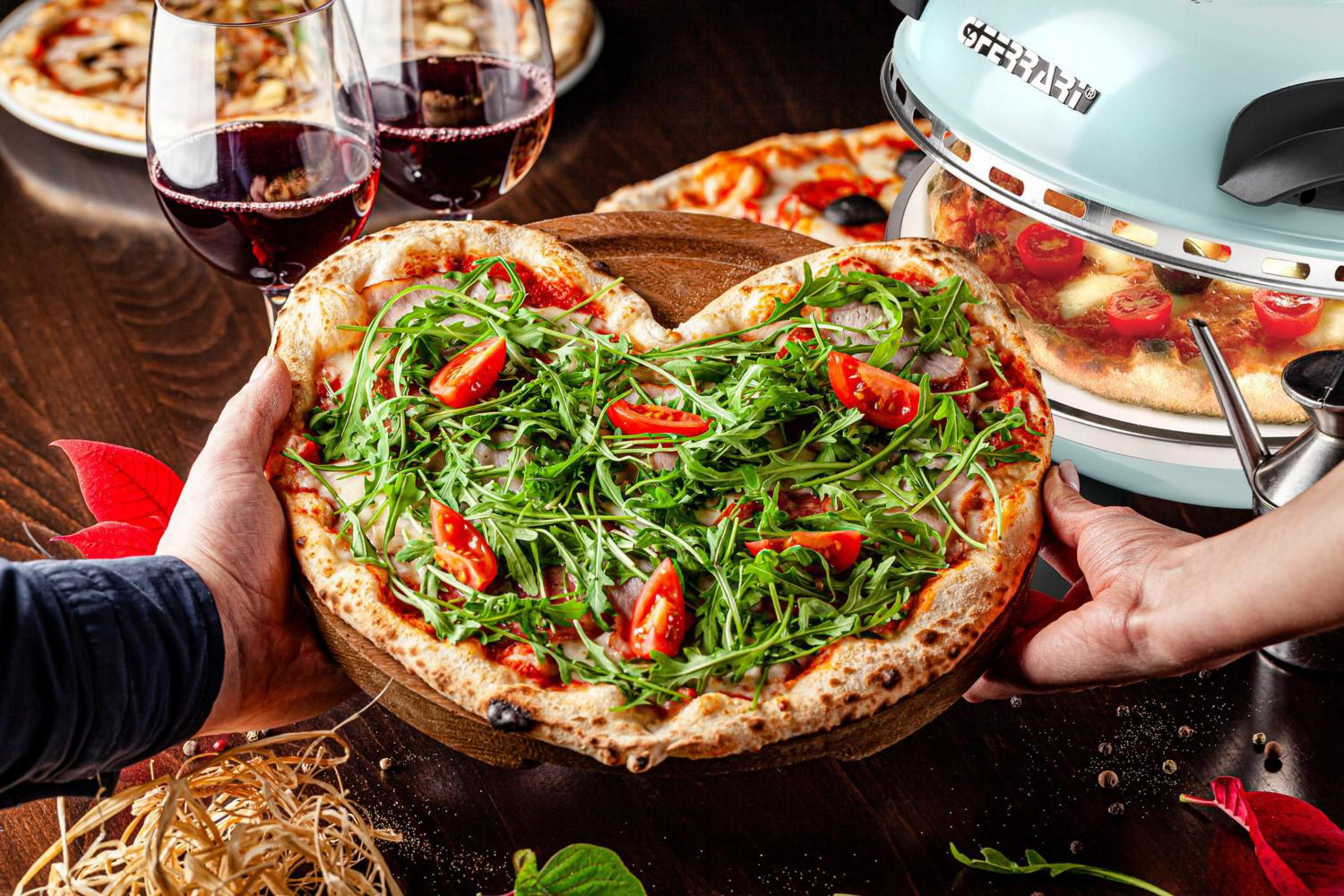 Pizza a forma di cuore su tagliere di legno, tenuta da due mani. Calici di vino rosso sullo sfondo.