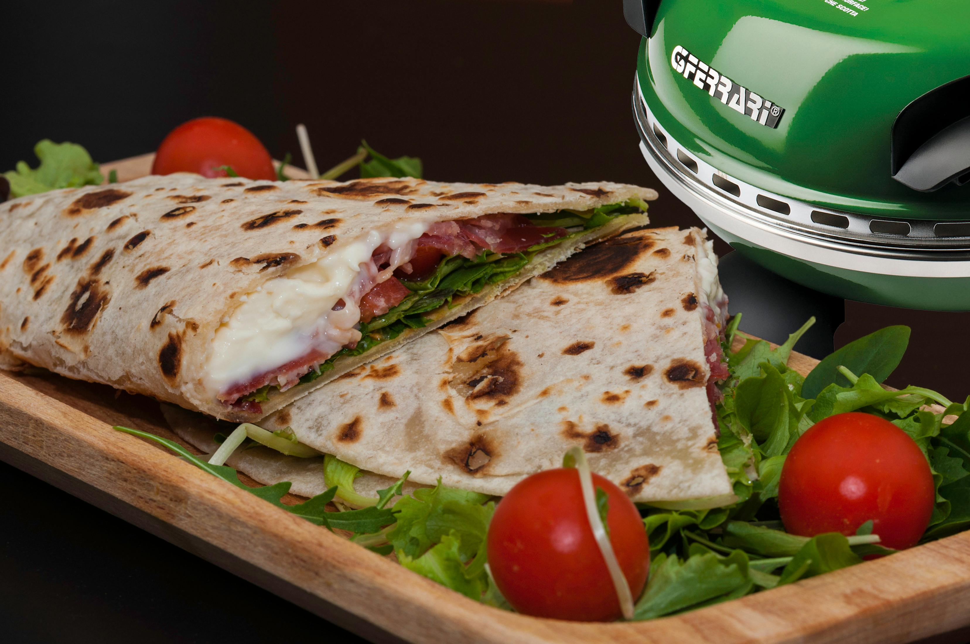 Panino piadina a metà su tagliere di legno con pomodori e lattuga, vicino a forno per pizza verde.