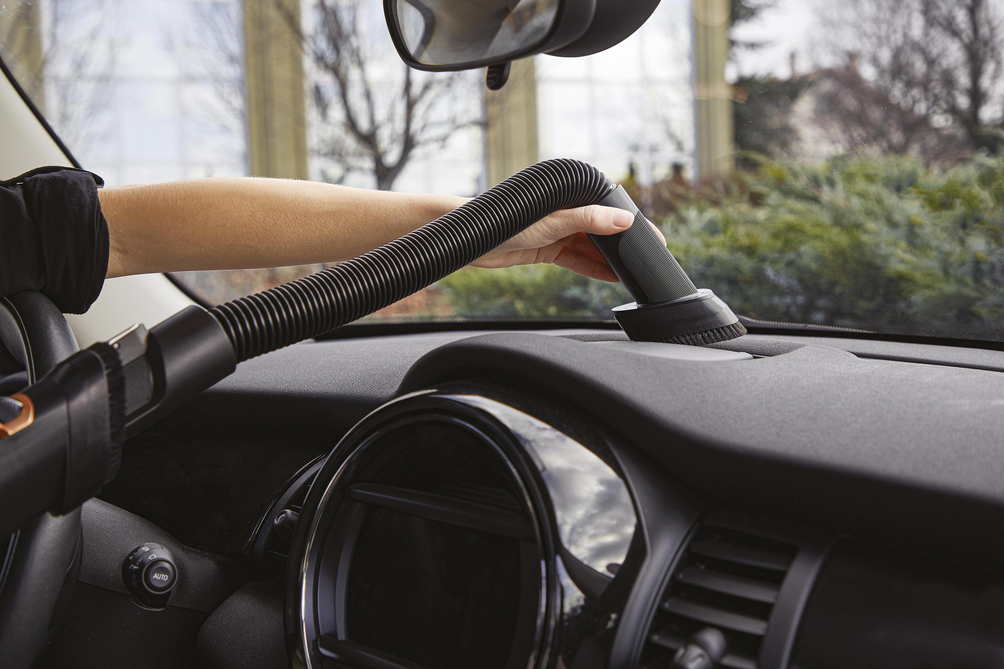 Osoba odkurza deskę rozdzielczą samochodu czarnym odkurzaczem z nasadką szczotkową.