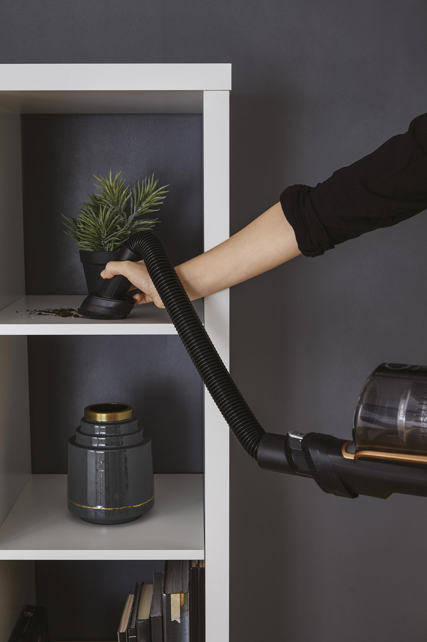 Ręka osoby używa odkurzacza do czyszczenia rośliny w białej szafce.