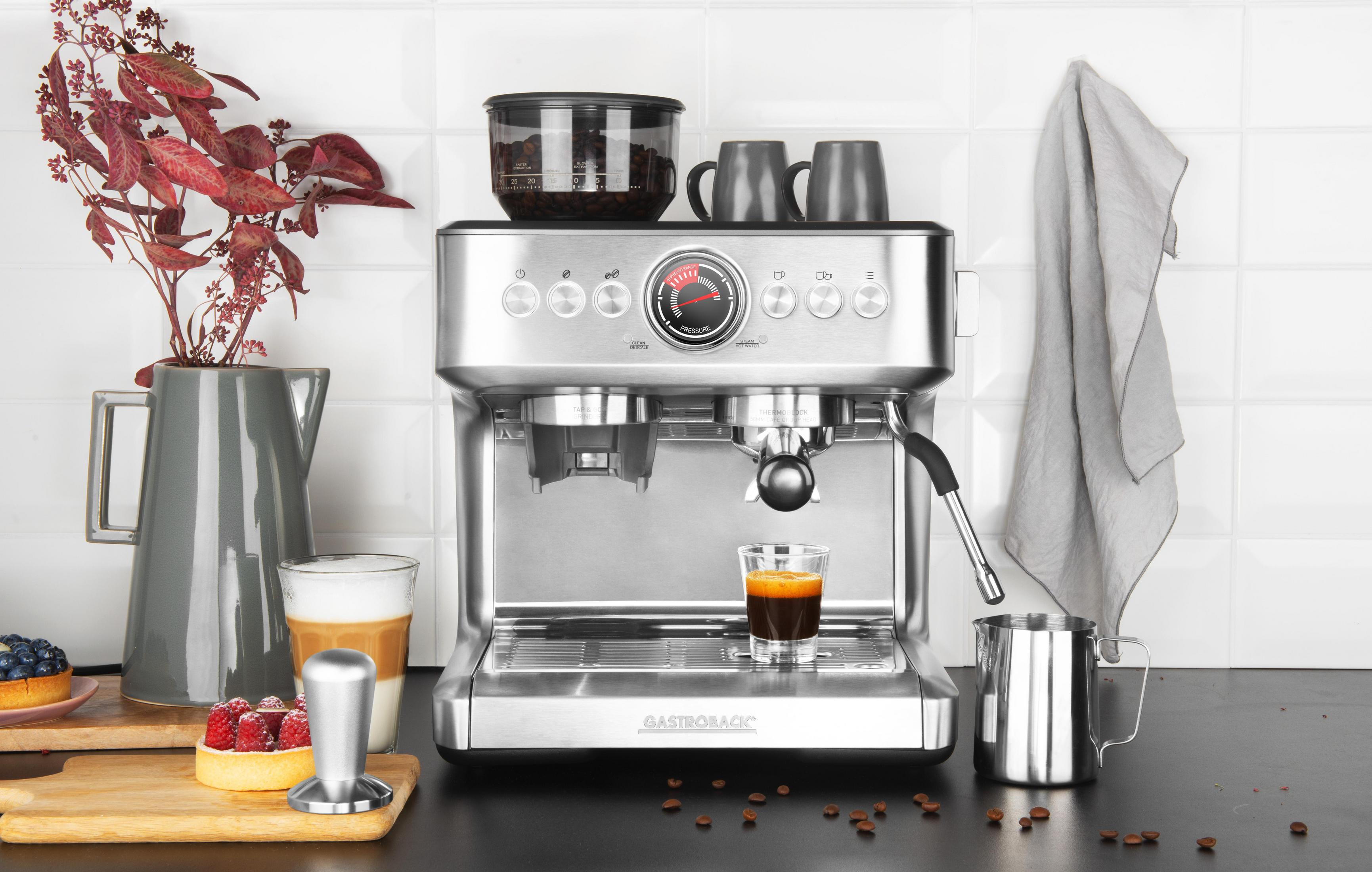 Una macchina per caffè espresso argentata con tazze, un bricco per il latte e chicchi di caffè. Bacche e dessert sul lato. Piastrelle bianche.