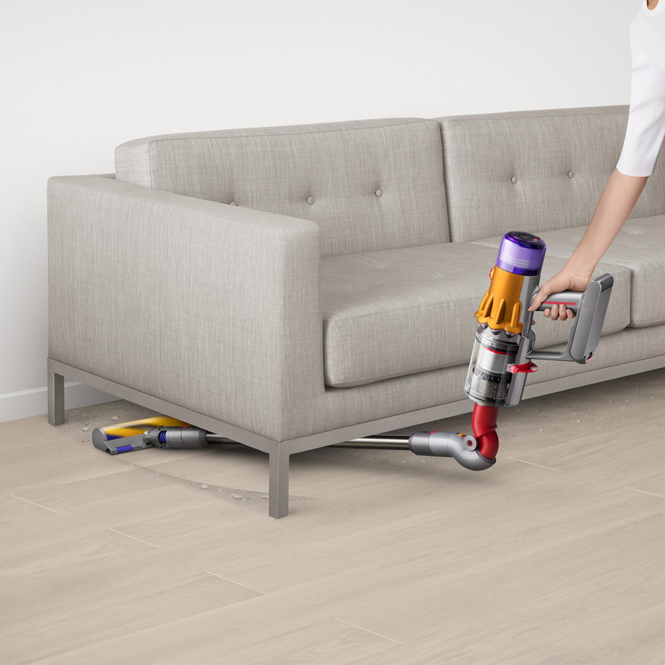 Une personne passe l'aspirateur sous un canapé gris avec un aspirateur Dyson. Sol beige.