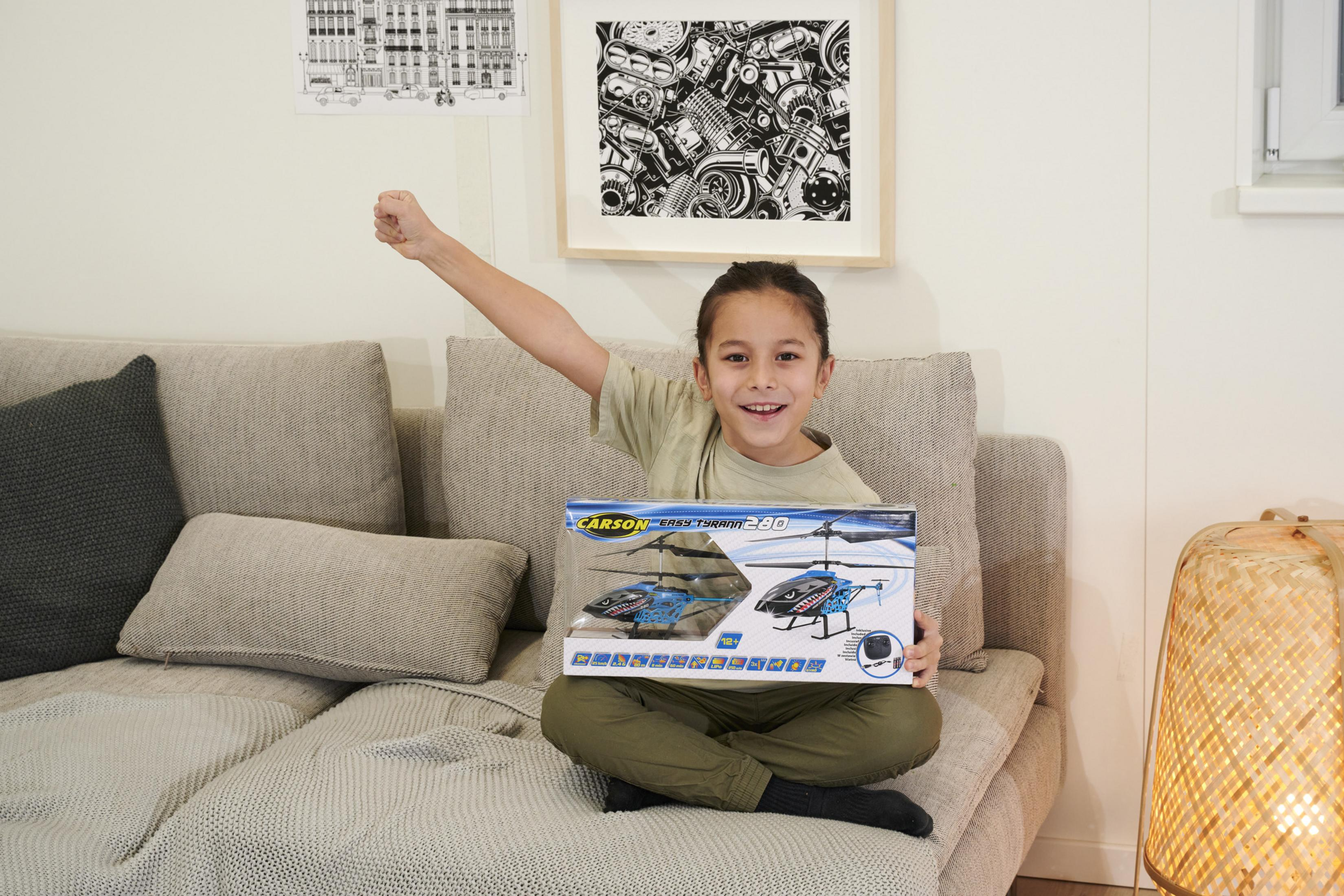 Ein Kind sitzt auf einer Couch, hält eine Spielzeug-Helikopter-Box und hebt einen Arm.