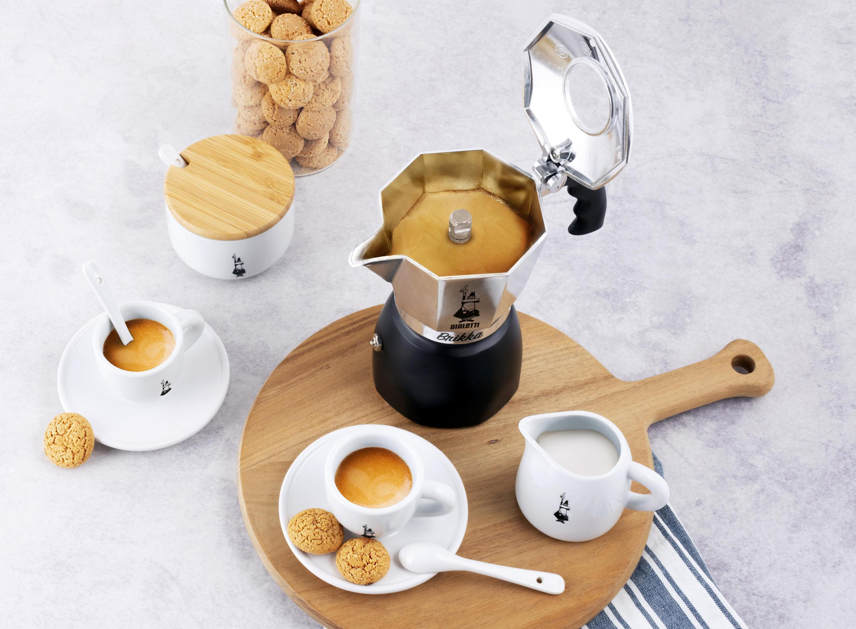 Kaffee-Setup: Moka-Kanne, Tassen, Milch und Kekse auf einem Holzbrett.