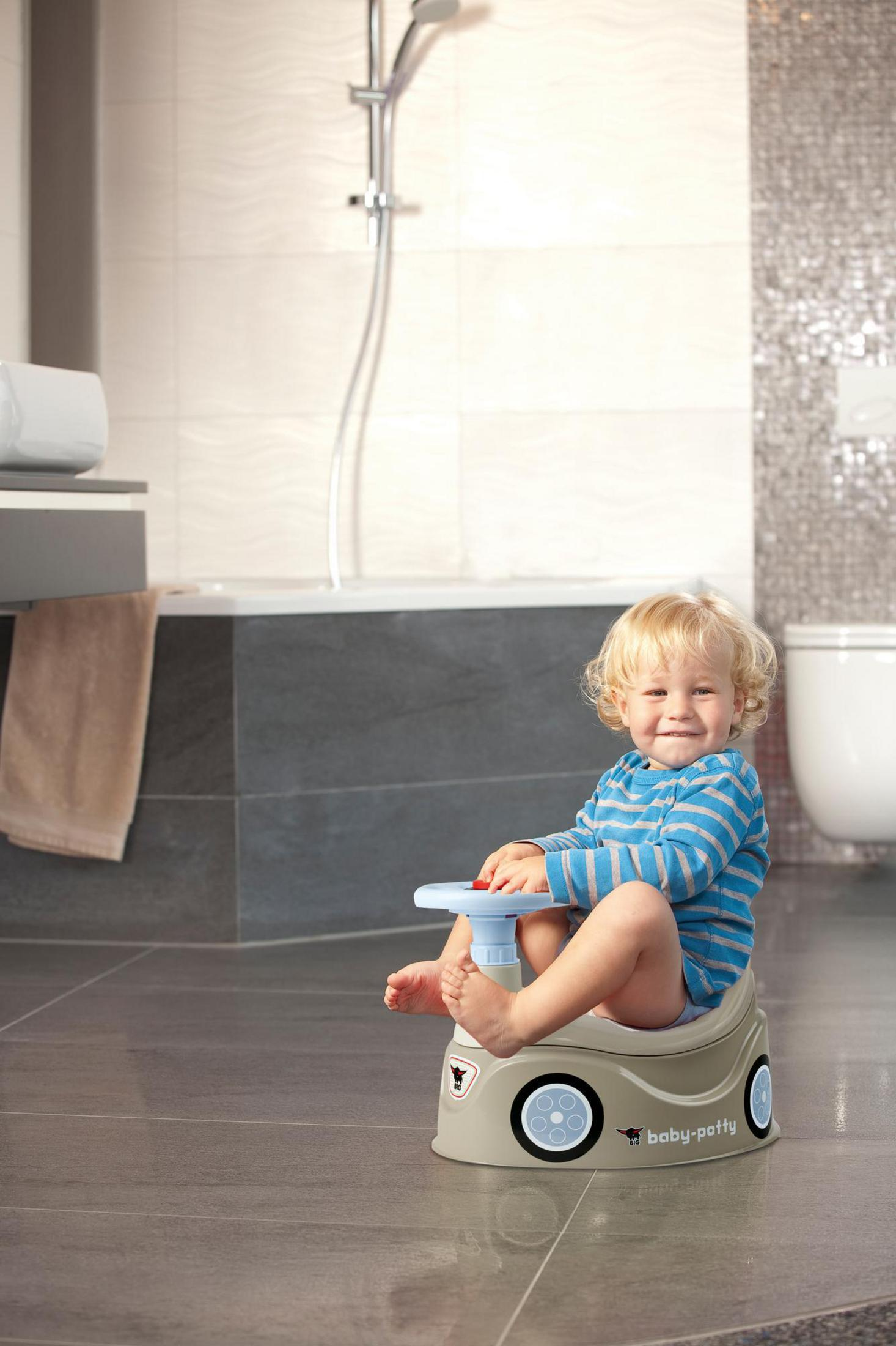 Kleinkind sitzt auf einem Baby-Töpfchen im Badezimmer, lächelt, weiße und graue Fliesen.