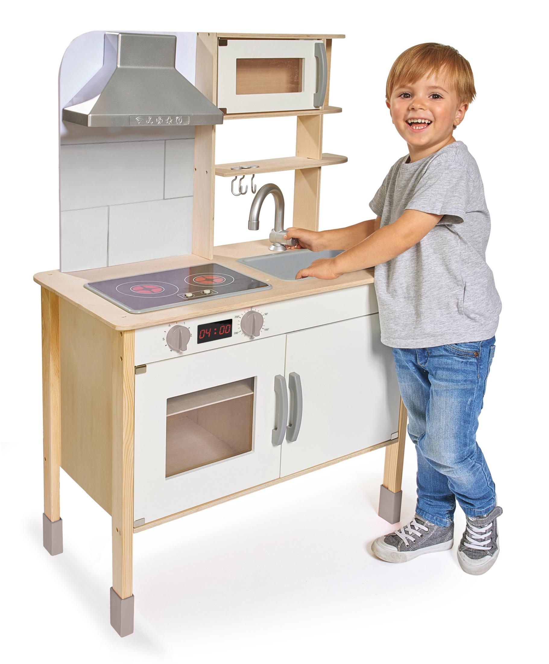 Un bambino sorride accanto a una cucina giocattolo in legno. Ha un fornello, un microonde, un lavandino e armadietti.