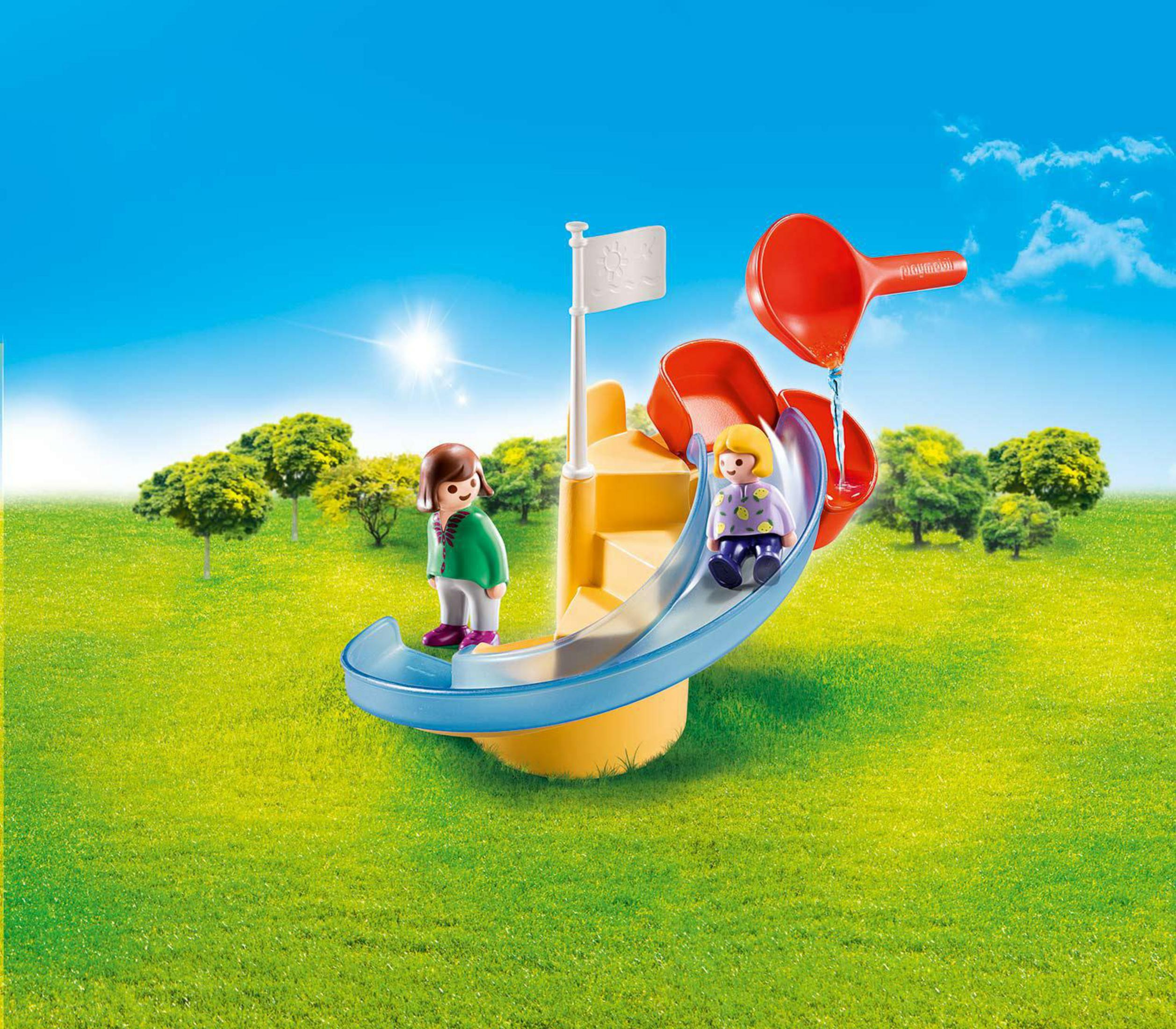 Zwei Figuren auf einer Rutsche, mit rotem Wasserspiel und grünem Gras im Hintergrund.