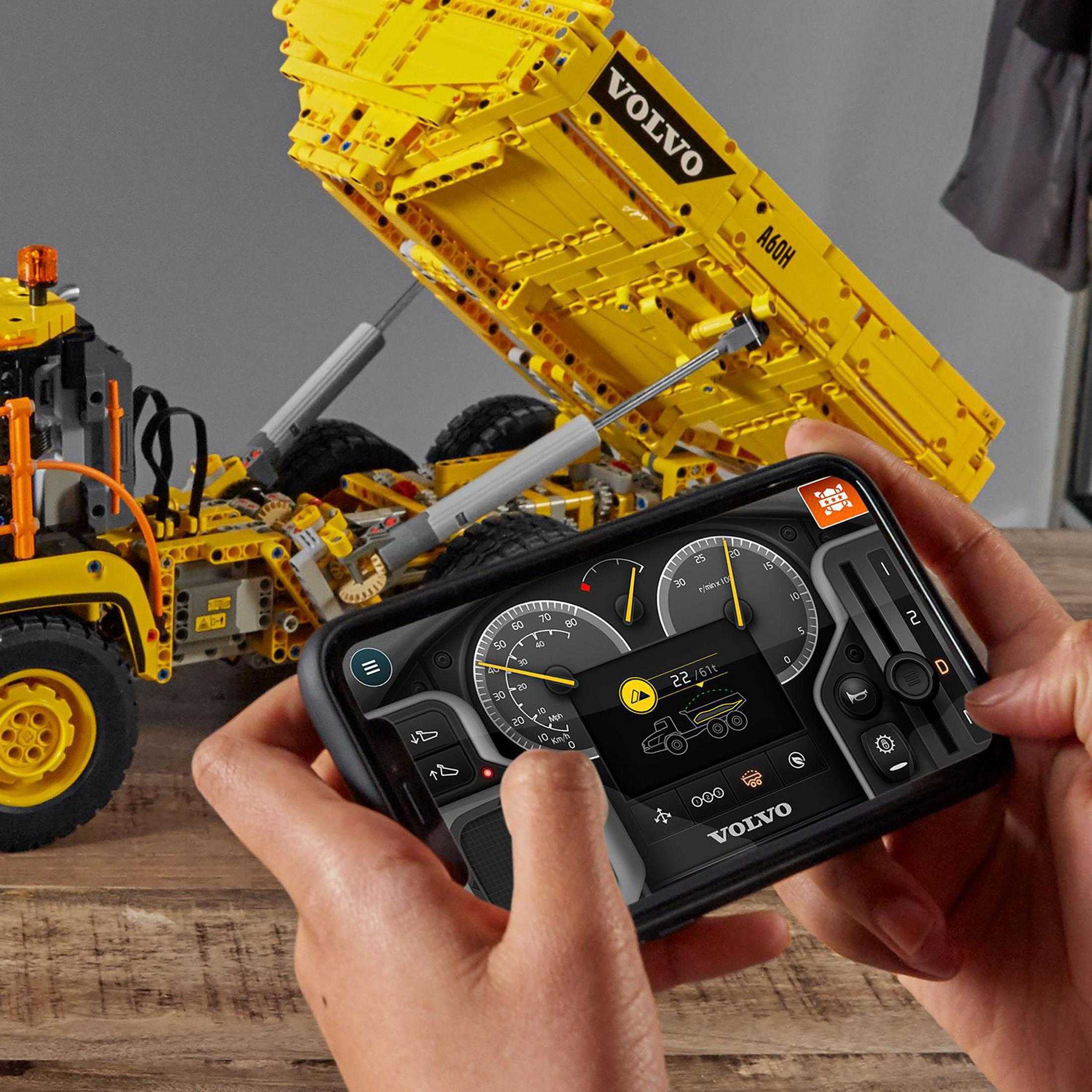 Una persona che tiene un telefono che controlla un camion Lego giallo su una superficie di legno.