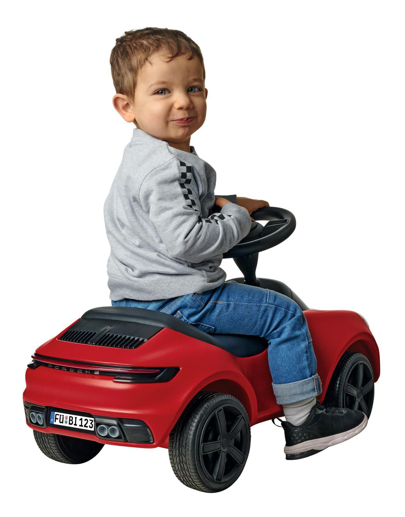 Ein kleiner Junge sitzt in einem roten Spielzeugauto und lächelt in die Kamera.