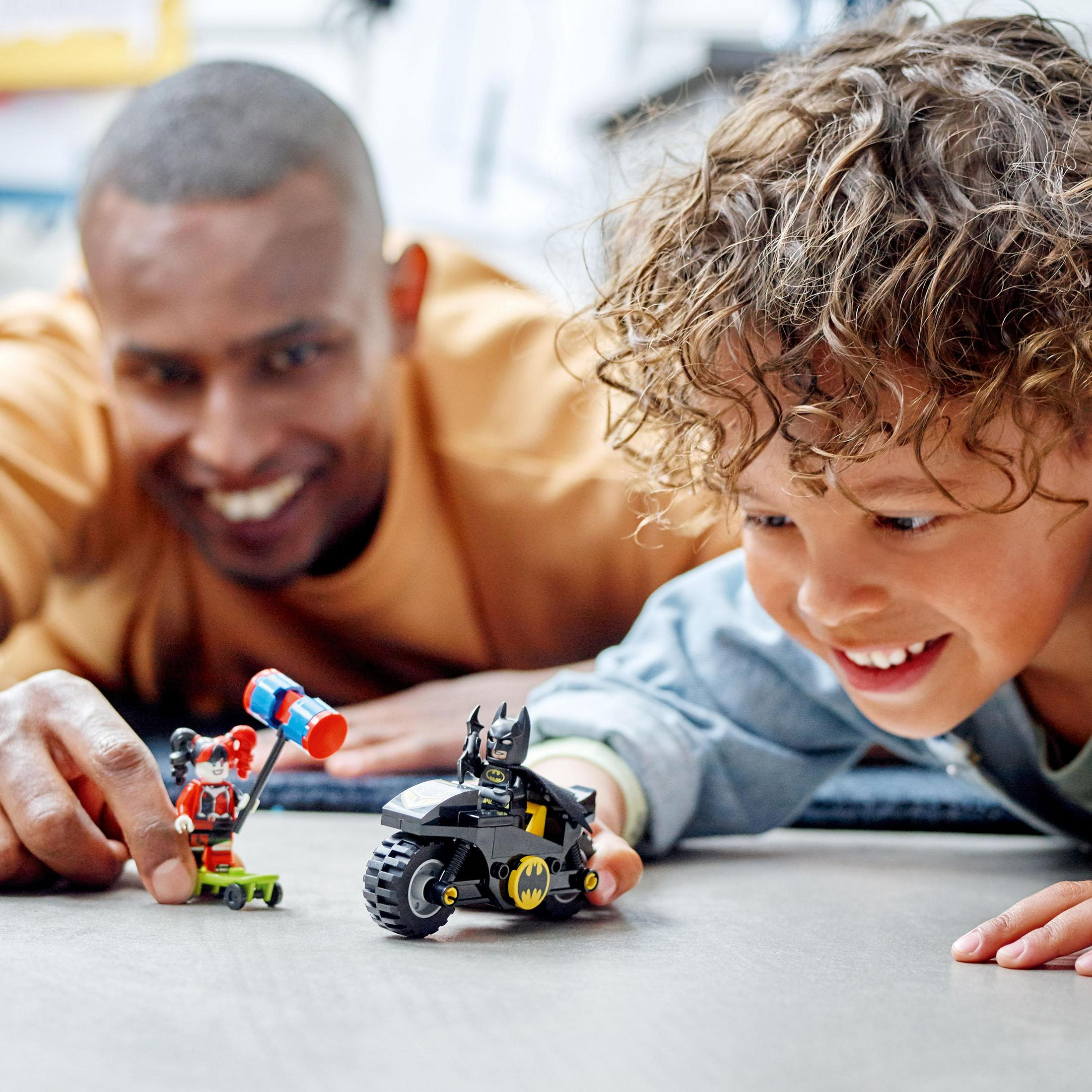 Ein Vater und Sohn spielen mit LEGO Batman- und Harley Quinn-Figuren auf einer grauen Oberfläche.