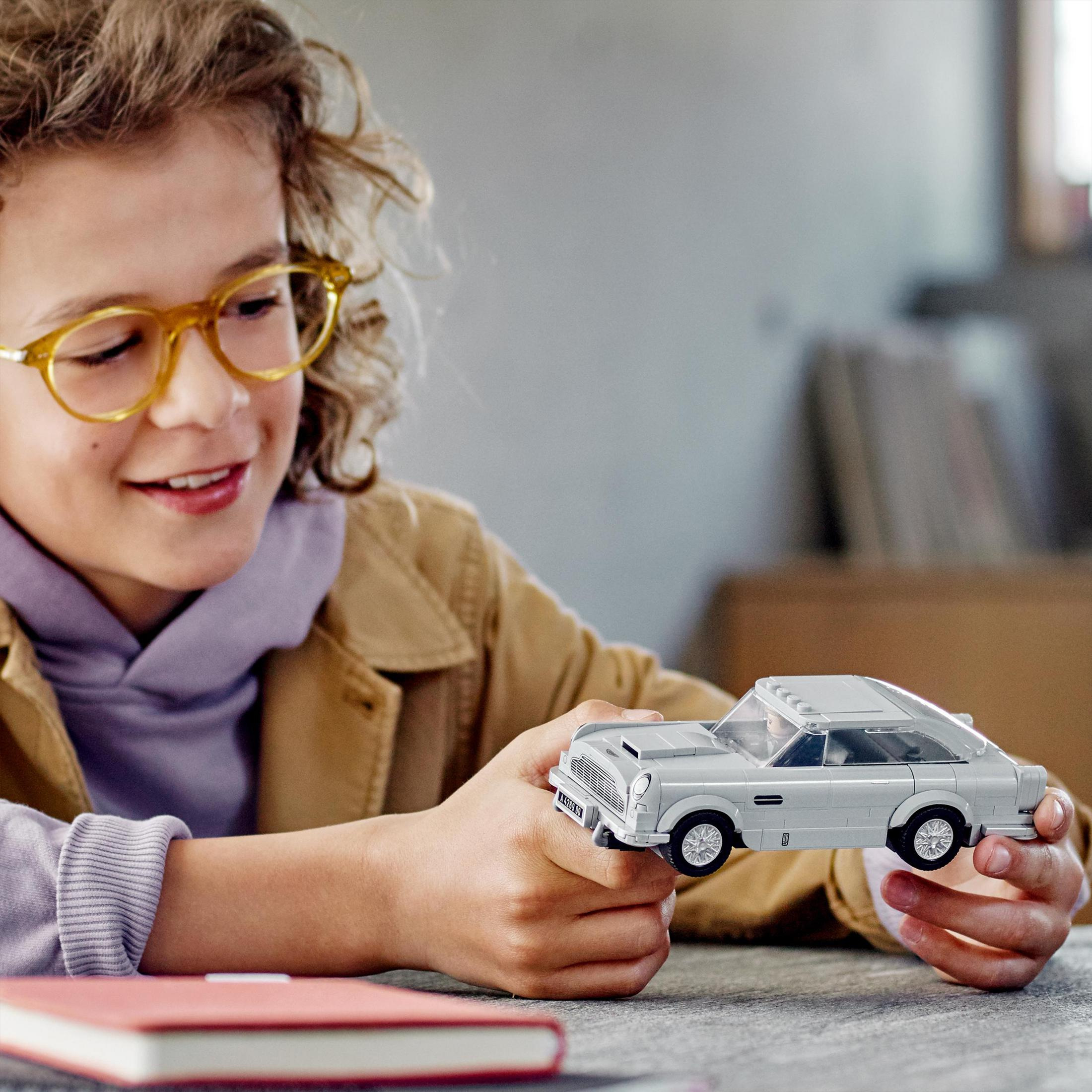 Ein Junge hält ein graues Spielzeugauto. Er lächelt. Das Auto ist ein klassisches Modell. Er trägt eine Brille und eine Jacke.