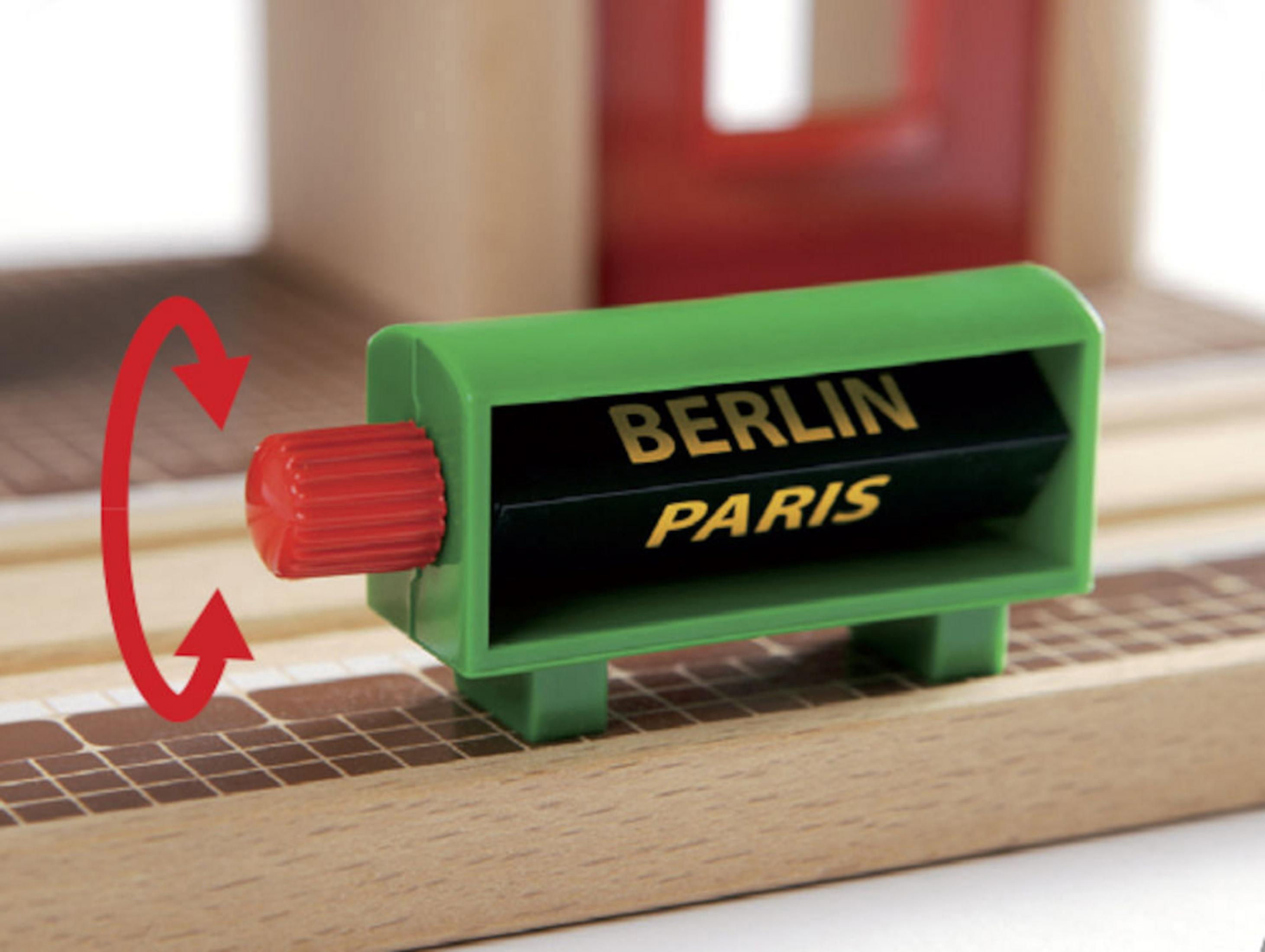 Grünes Bahnhofsschild mit Berlin und Paris. Roter Drehknopf zeigt die Richtung an.