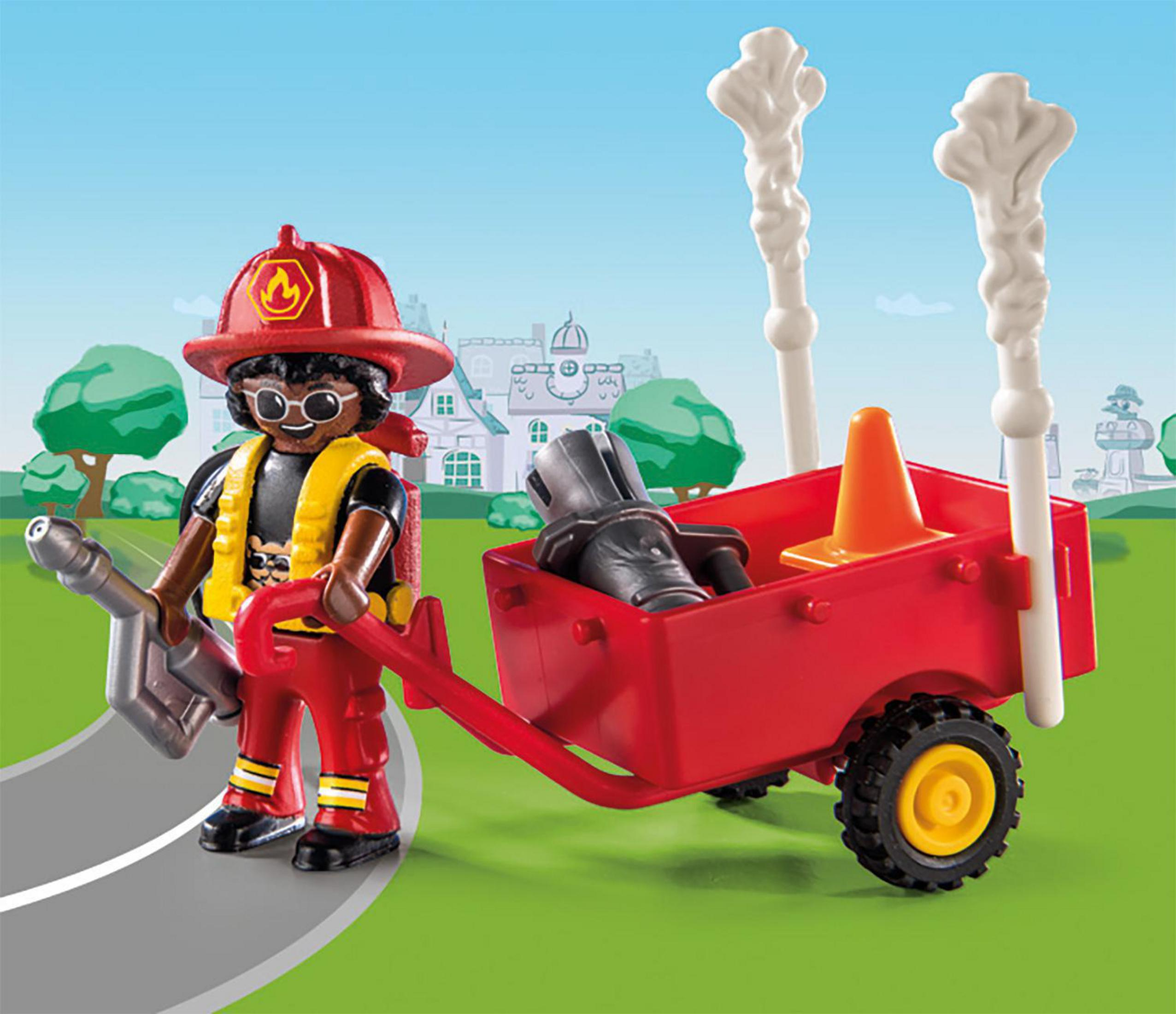 Ein Playmobil-Feuerwehrmann mit rotem Wagen. Er steht auf einer Straße mit Gebäudehintergrund.