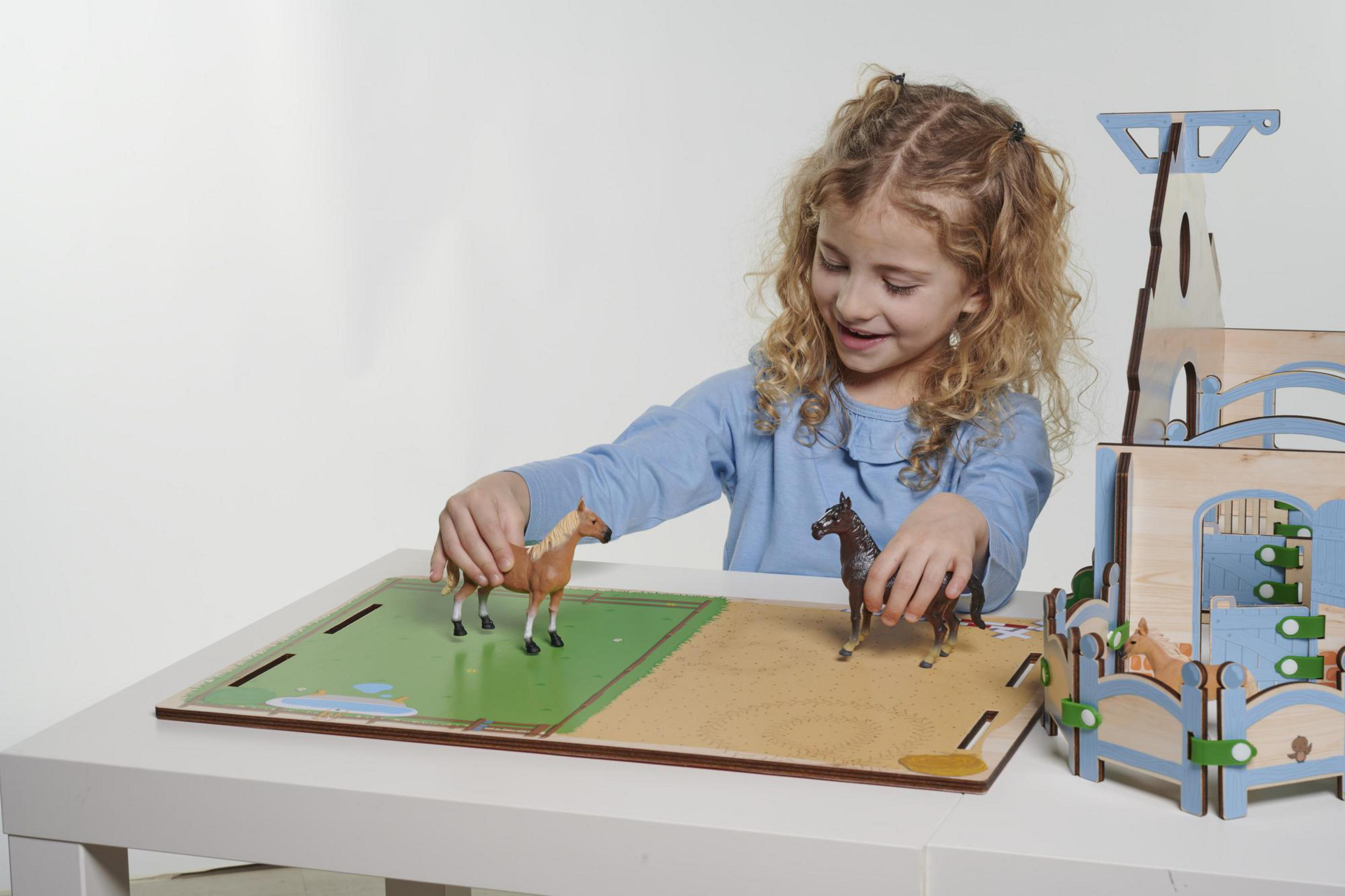 Una bambina gioca con cavalli giocattolo su un set di gioco in legno, sorridendo.
