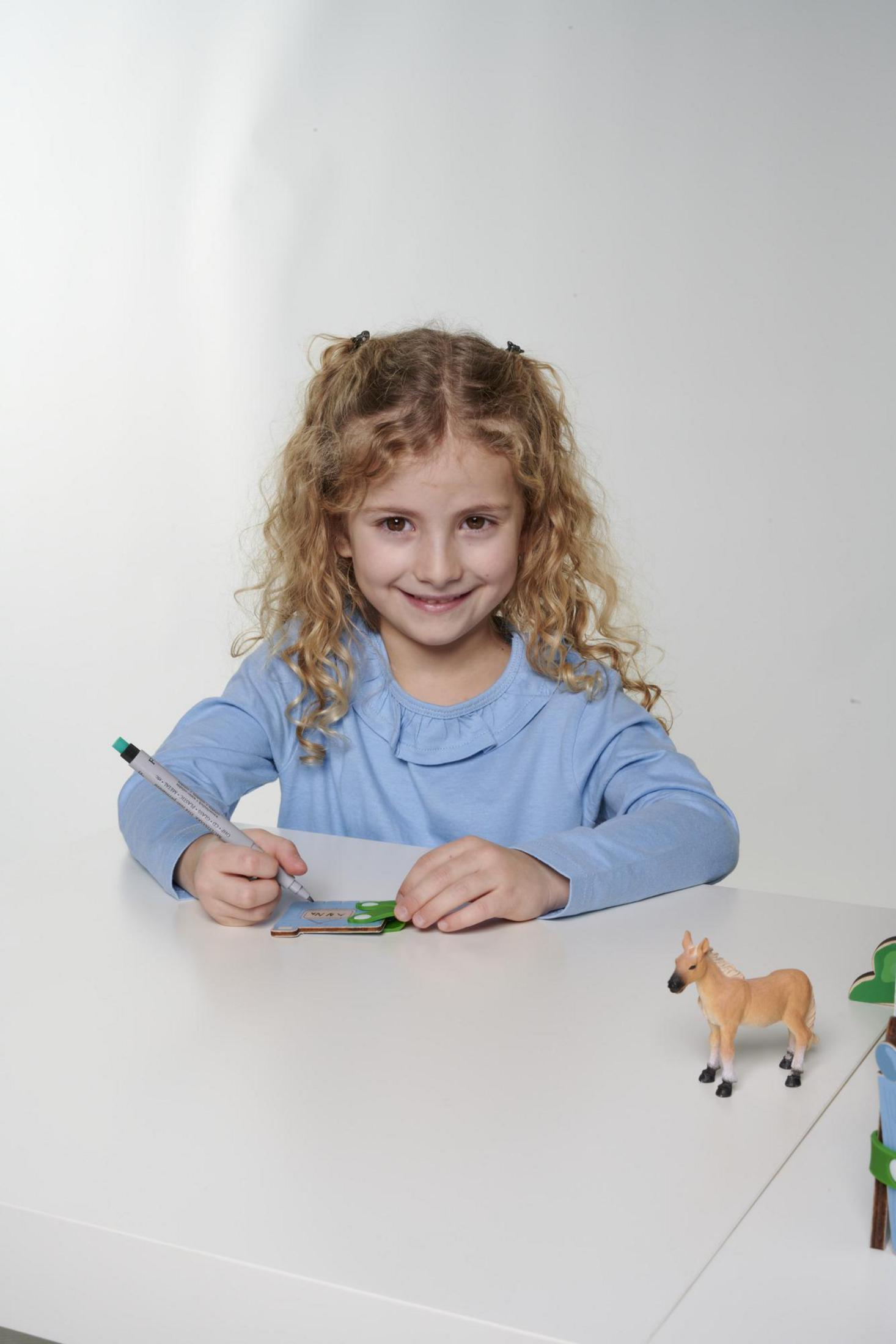 Una ragazza con i capelli ricci disegna su una carta. Un cavallo giocattolo è sul tavolo.
