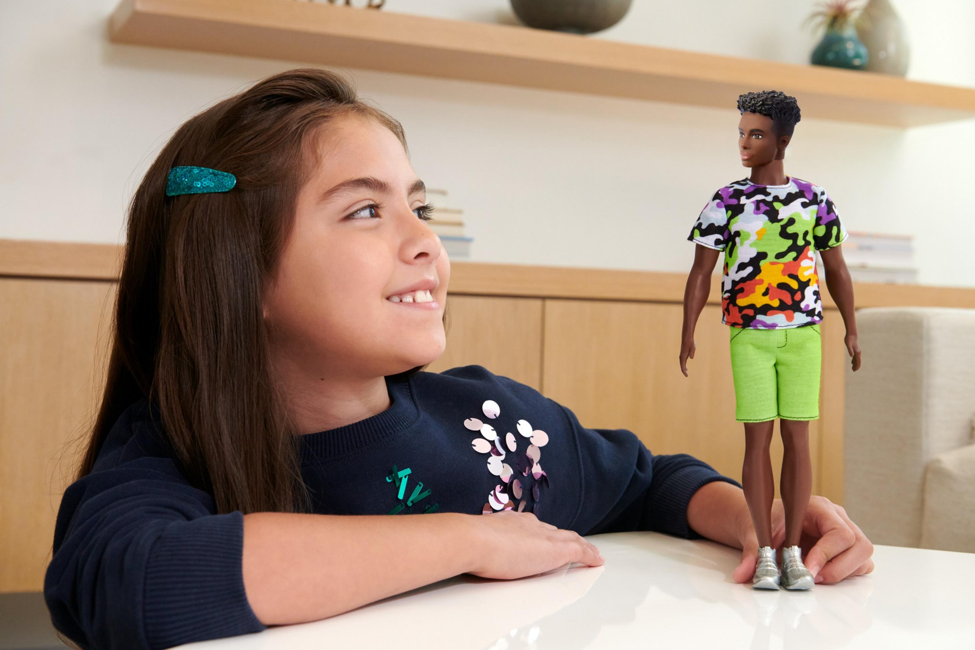 Dziewczynka uśmiecha się, patrząc na lalkę. Lalka to czarnoskóry Ken w kolorowym t-shircie i spodenkach.