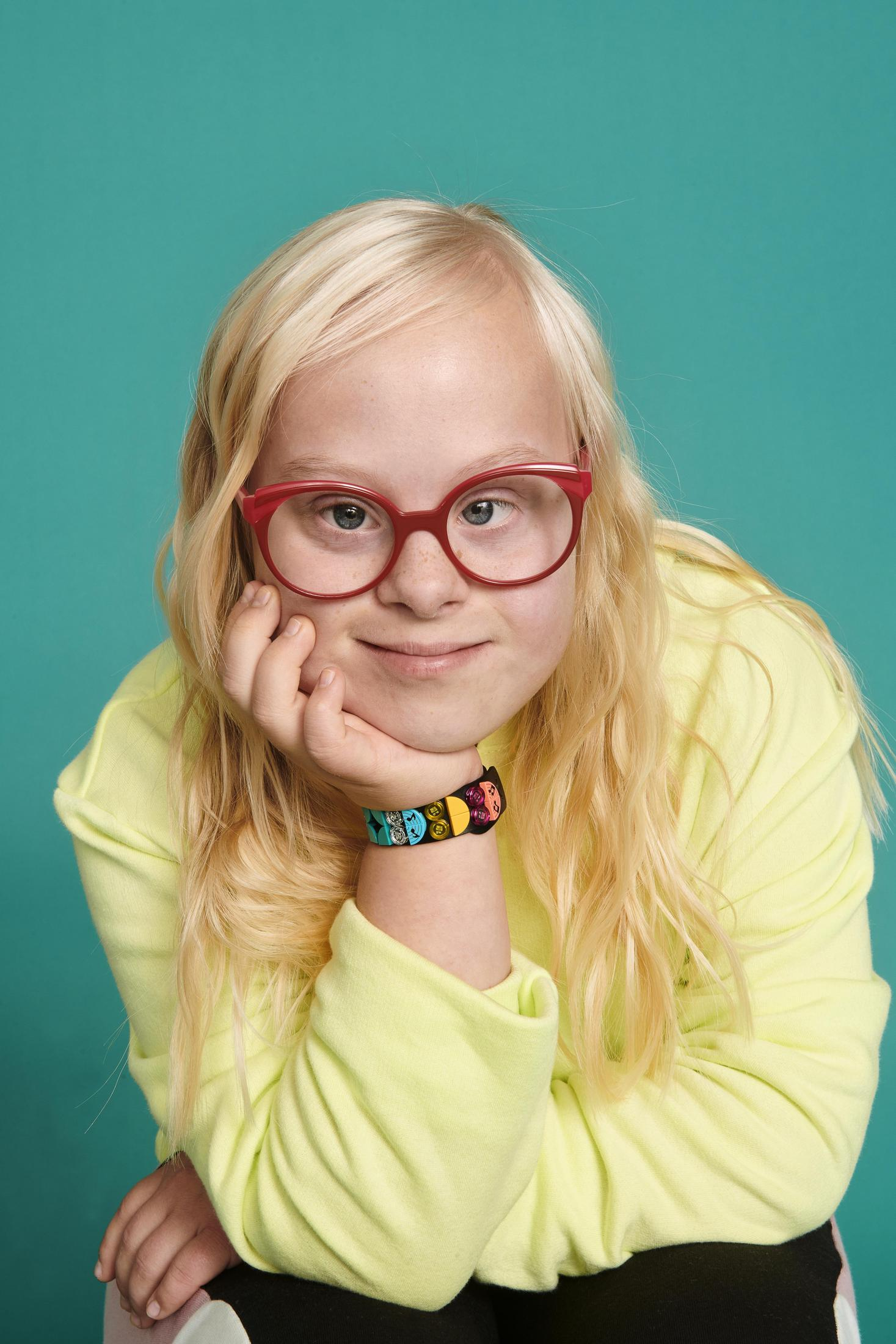 Blondynka w okularach uśmiecha się, opierając podbródek na dłoni.