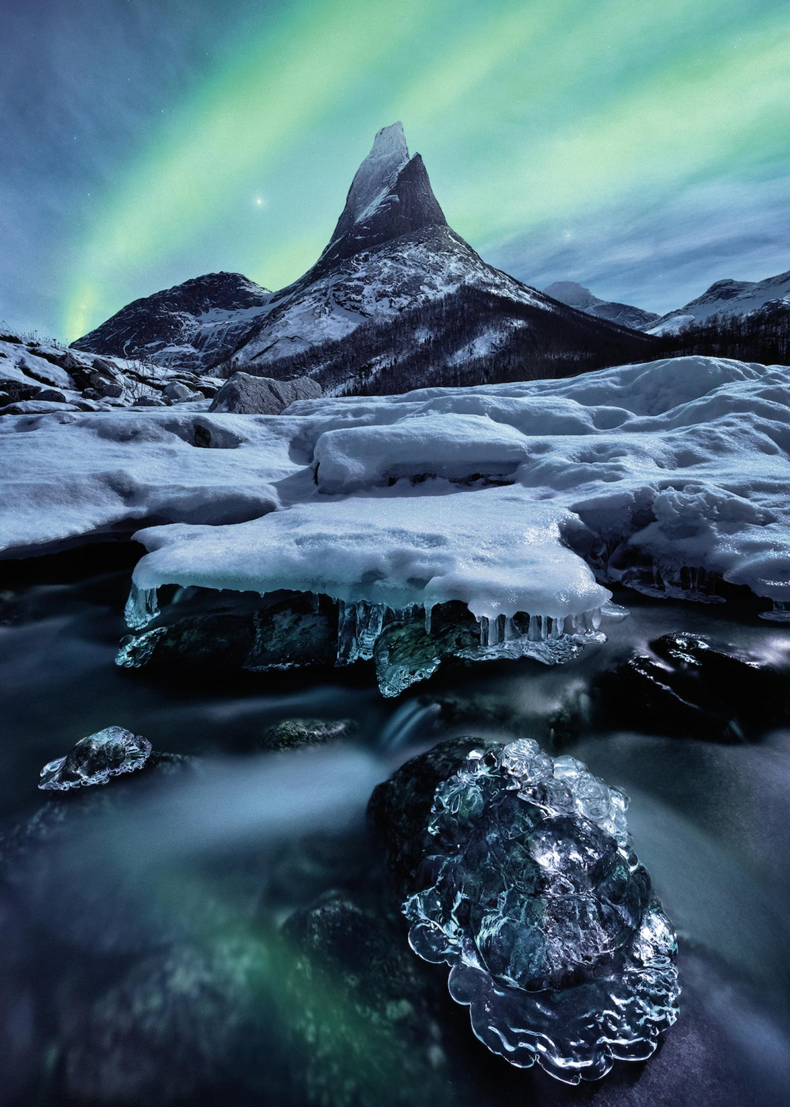 Eine Winterlandschaft mit Berg, Fluss, Schnee und Eis. Der Himmel hat grüne Polarlichter.