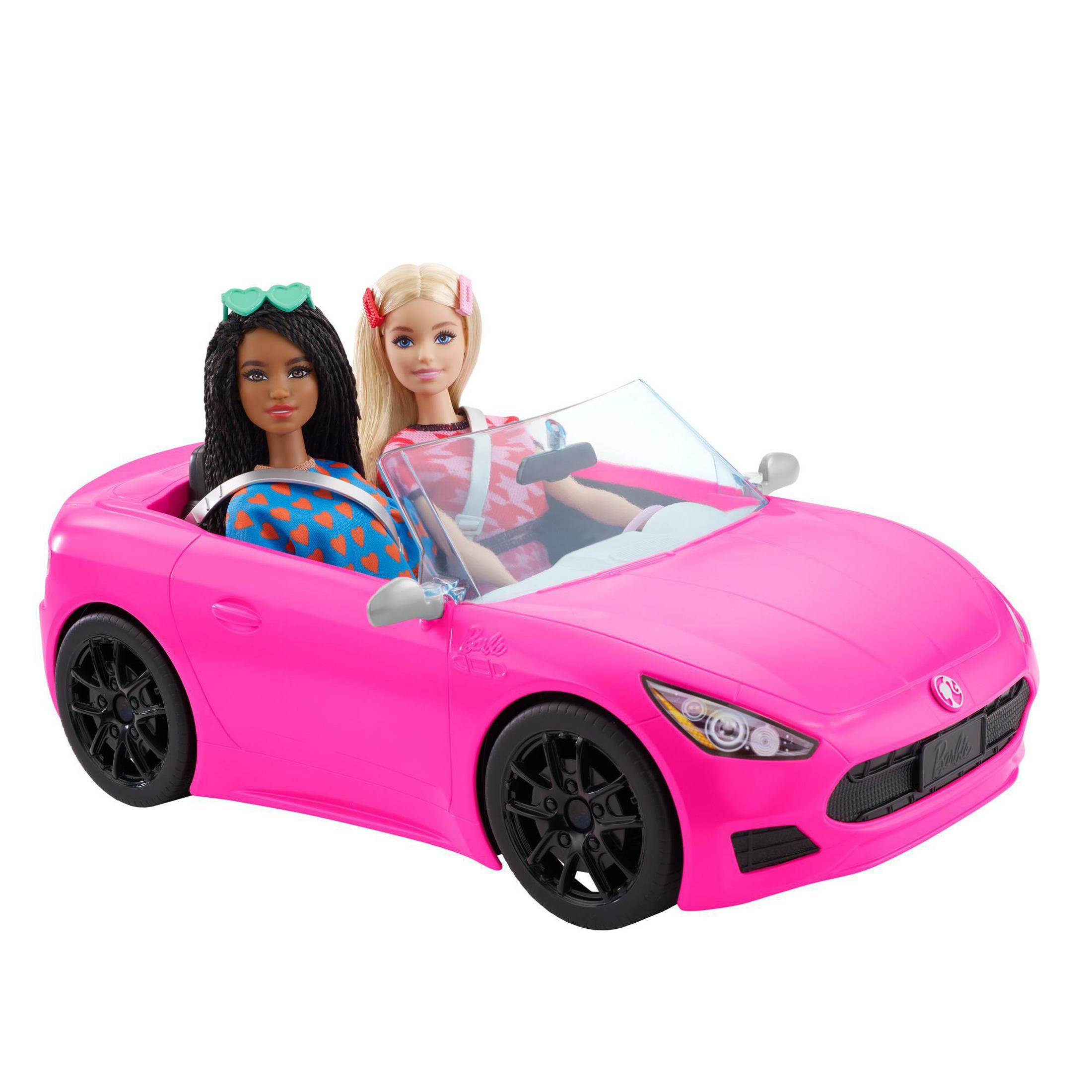 Zwei Barbie-Puppen in einem pinkfarbenen Cabrio. Eine Puppe hat dunkles Haar, die andere blond.