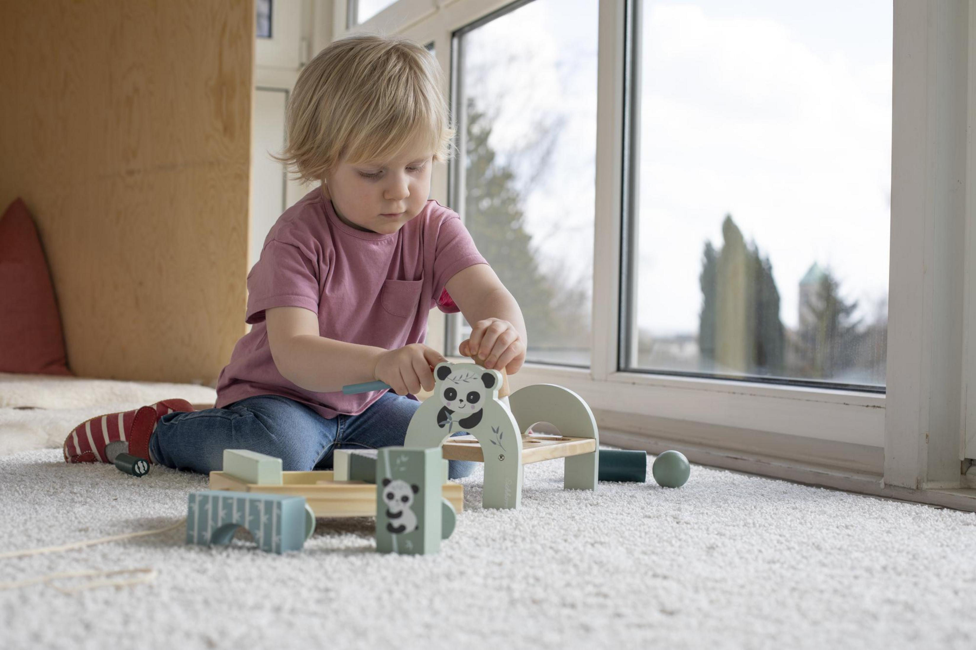 Kleinkind spielt mit hölzernen Pandablocks. Teppich und Fenster im Hintergrund.